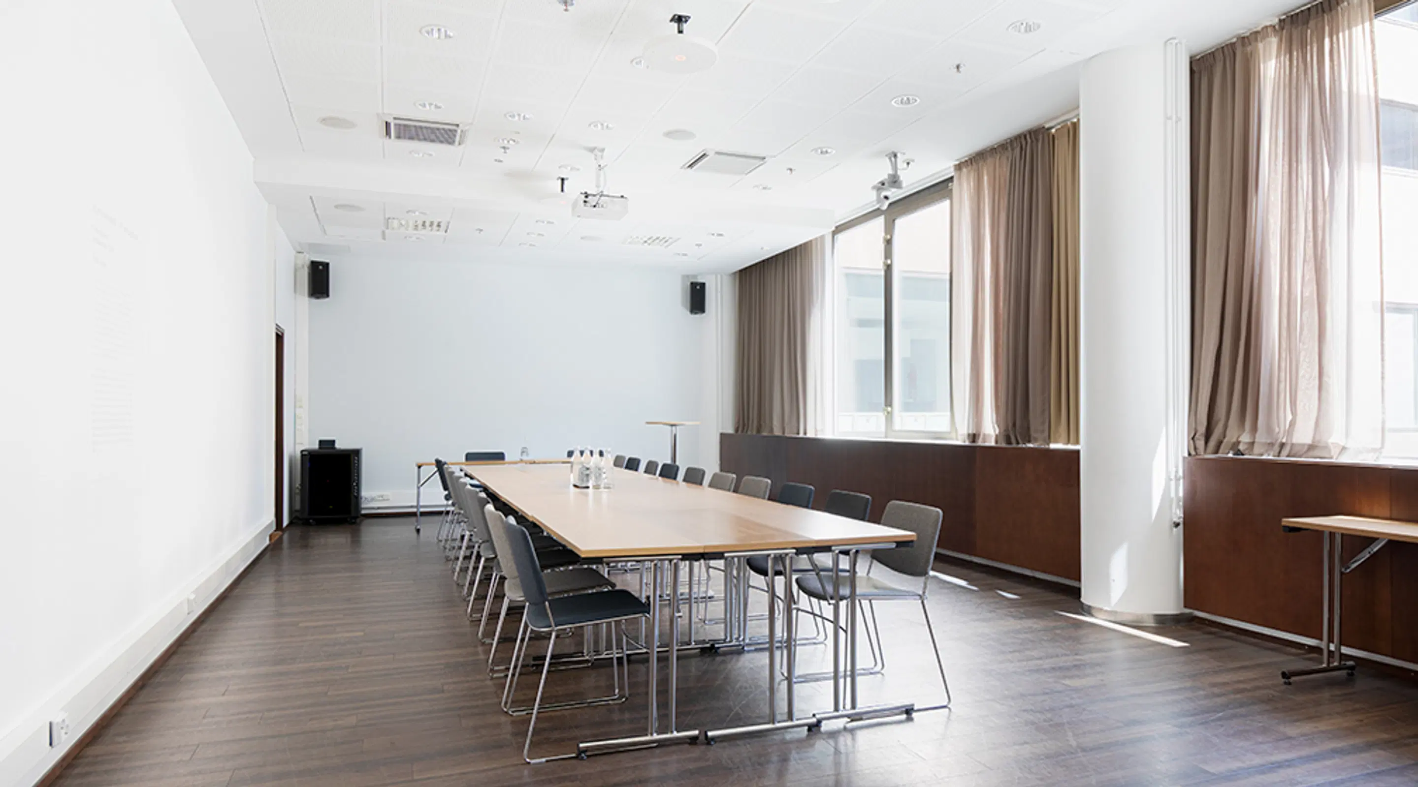 Bright meeting room with a long table and chairs, dark wooden floor and large windows with beige curtains.