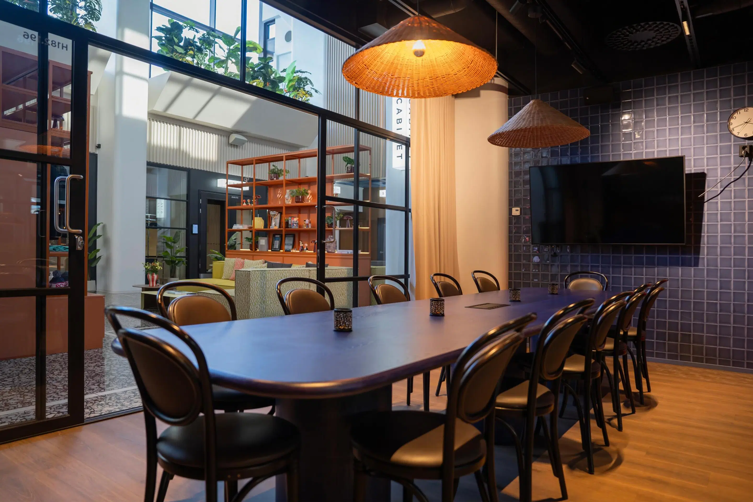 Modern meeting room with long table and chairs, hanging rattan lamps, and wall-mounted screen. Glass walls, wooden flooring, and shelving units in adjoining space.