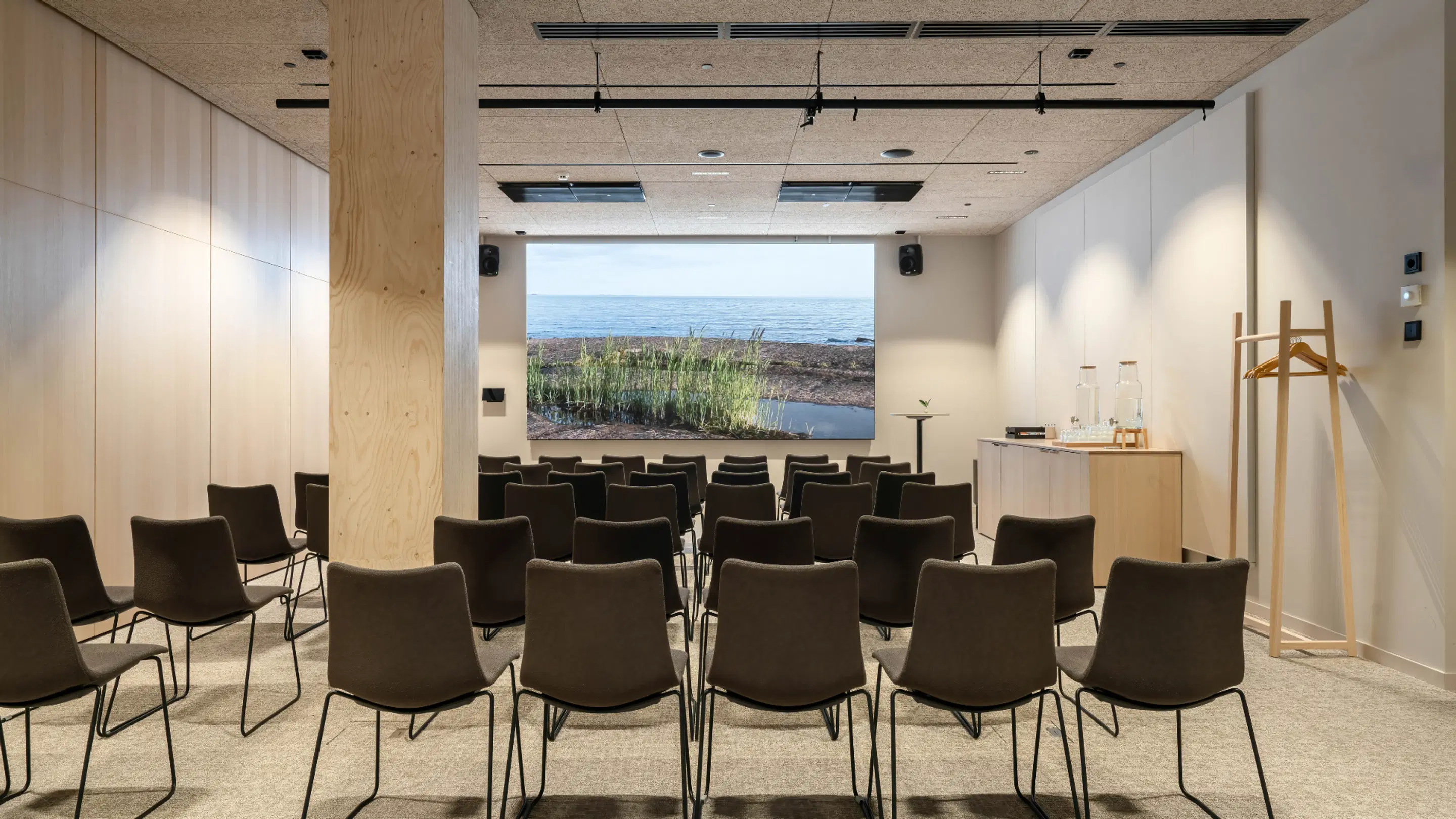 Modern auditorium with rows of dark chairs, large nature image on screen and light wooden wall paneling.