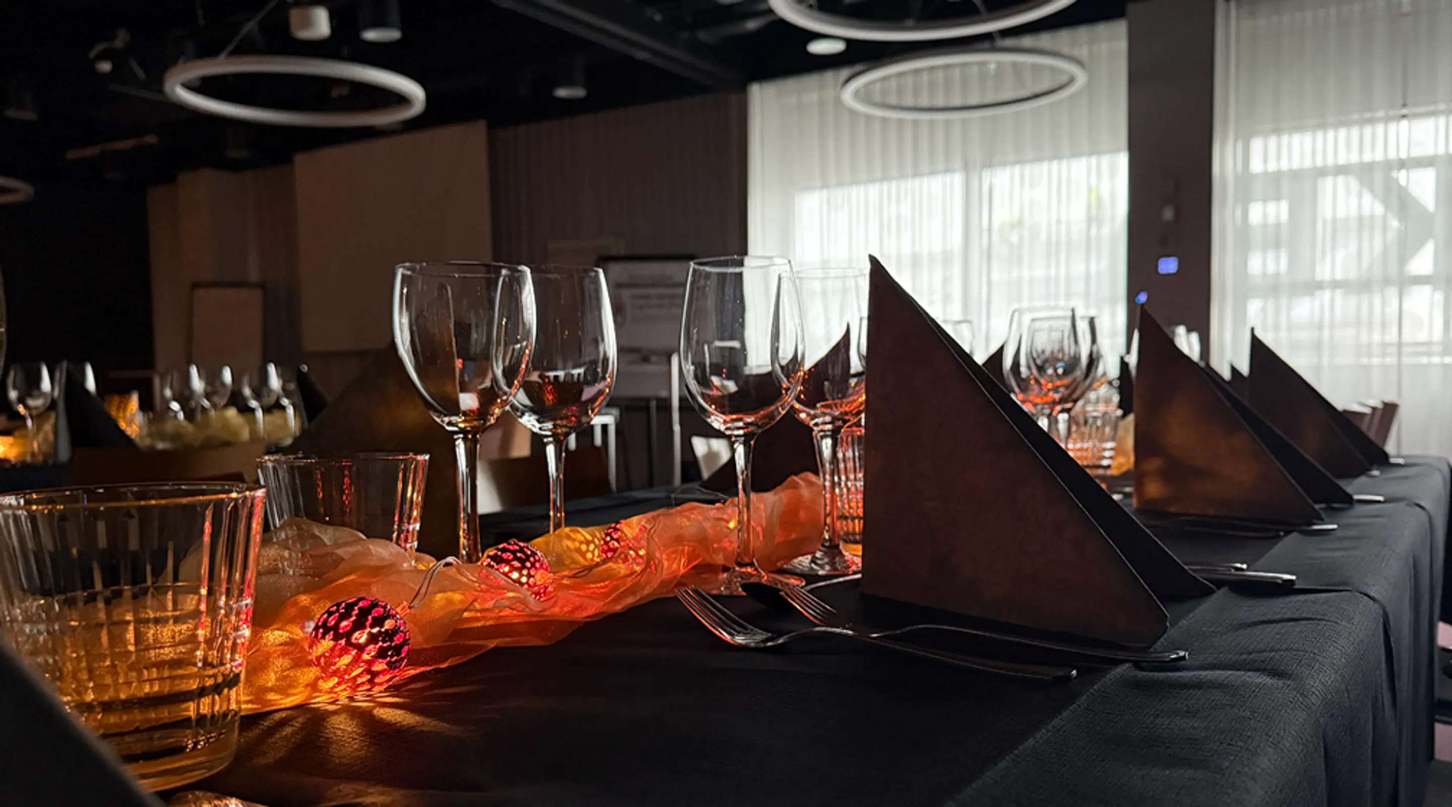 A table covered with dark cloth, set with wine glasses, folded napkins and decorative lights. The room has large windows and ring-shaped ceiling lights.