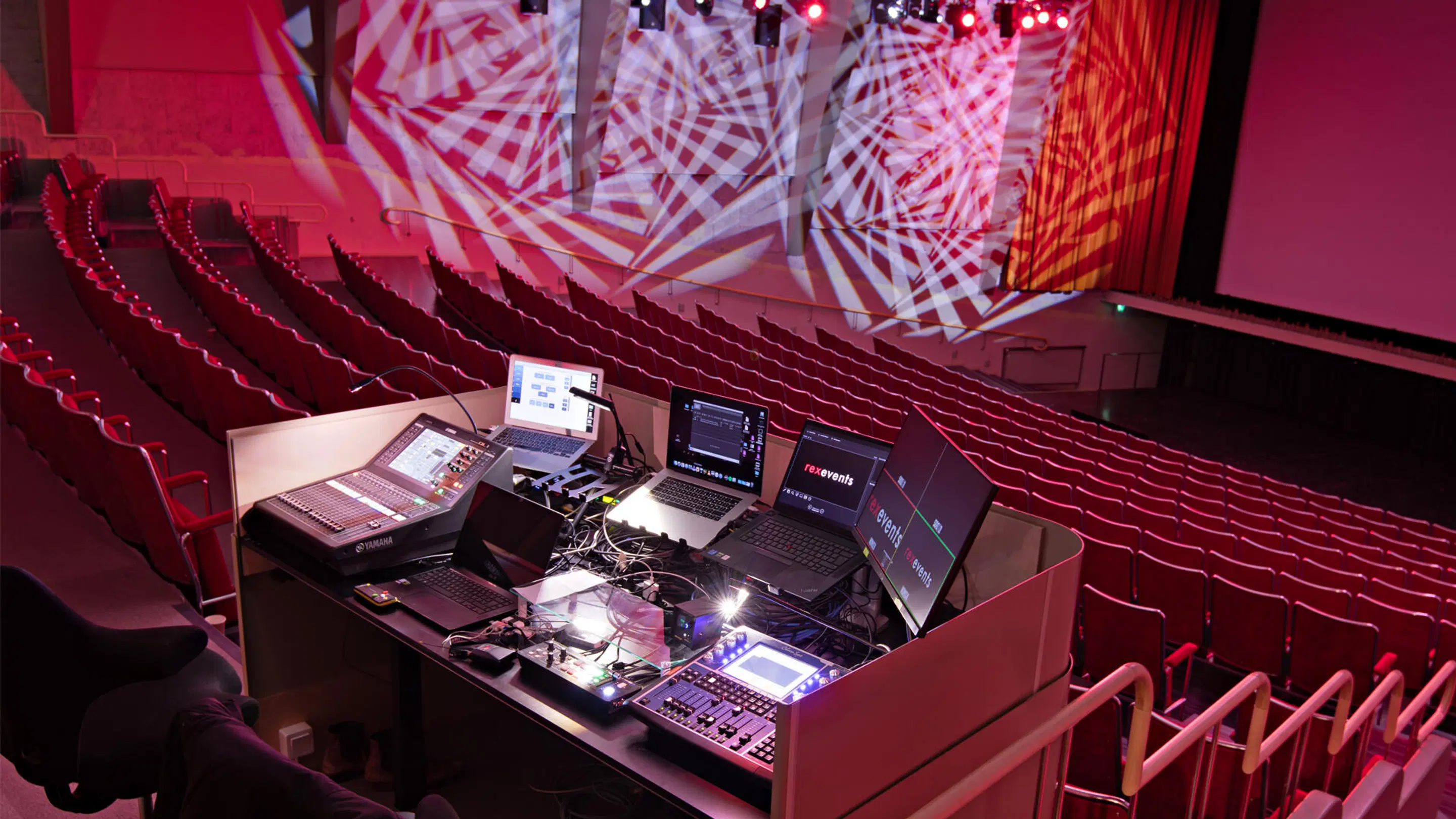 Technical desk with audiovisual equipment in an auditorium. Red seats and illuminated screen and stage under red lighting.