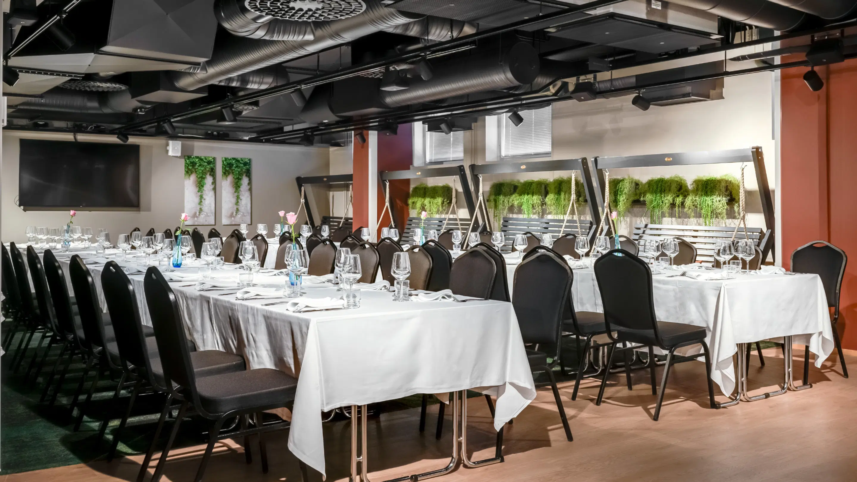 Modern meeting room with long tables and chairs. White tablecloths, table settings and wine glasses. Green plant elements in decor and a display screen on wall. Hard floor surface.