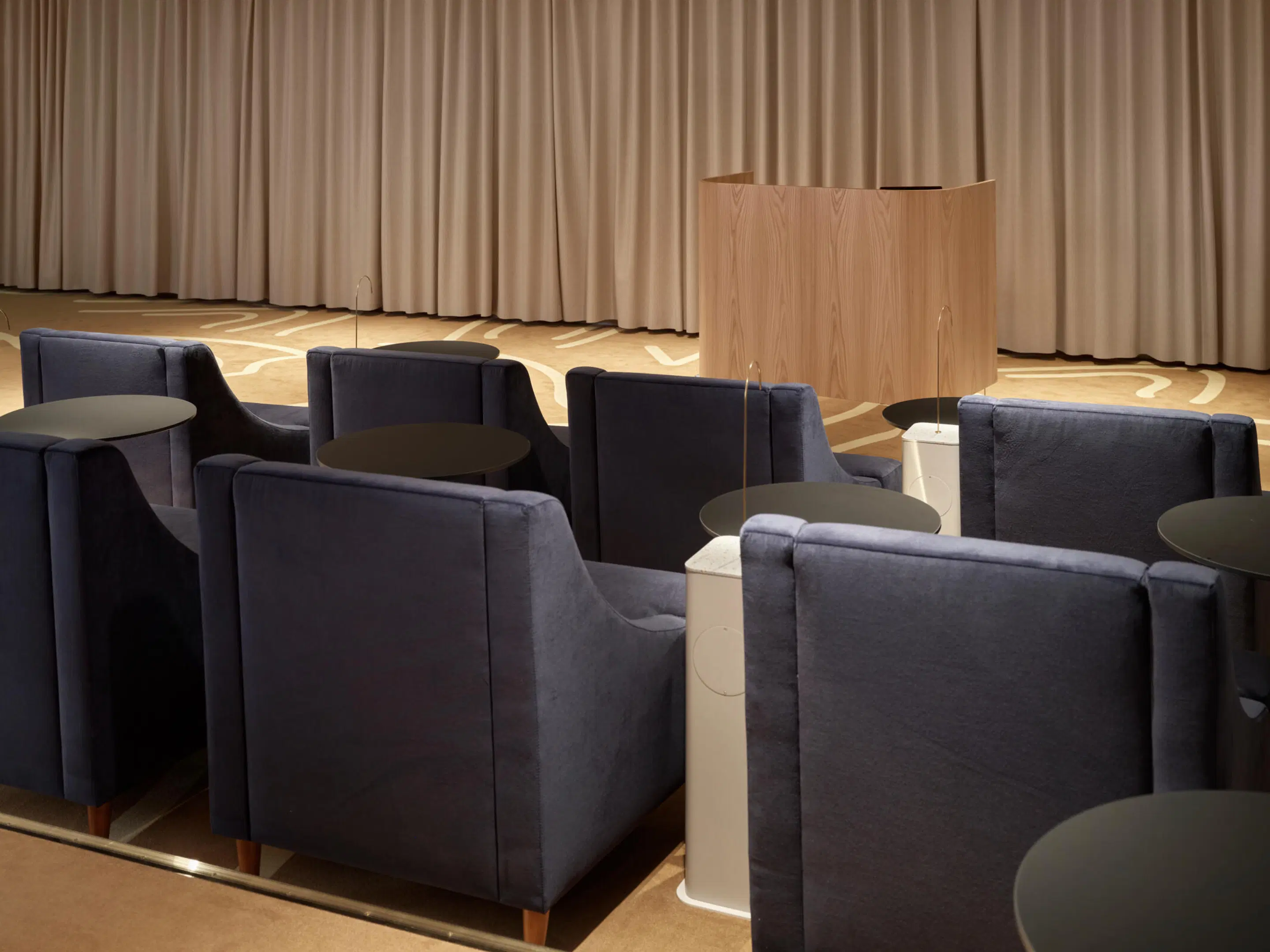 Auditorium med mörkblå fåtöljer och små bord. Framför finns en träpodium och beige gardiner i bakgrunden. Golvet är av träparkett.