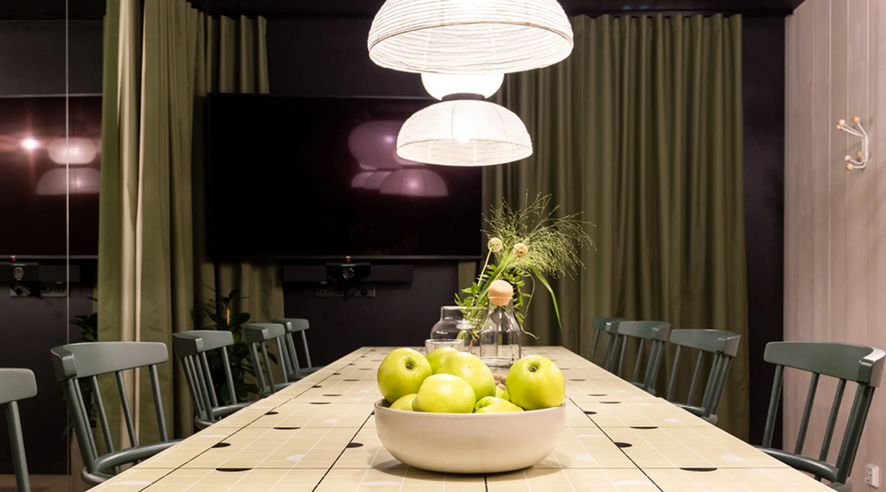 Wooden dining table with green chairs, a bowl of green apples and a plant vase. Two dome-shaped pendant lights hang above the table.