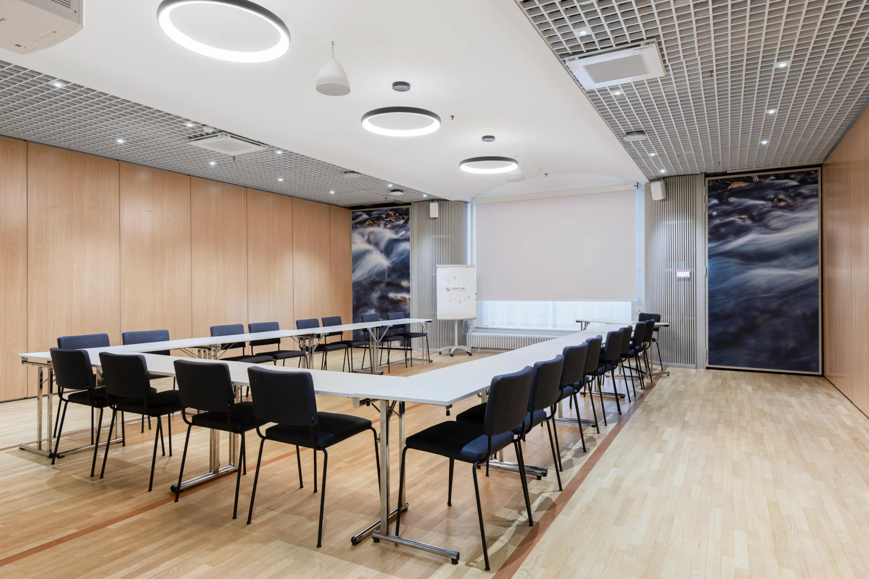 Modern meeting room with U-shaped table arrangement, wooden panel walls, water-themed artwork and level wooden floor. Bright circular LED lighting overhead.