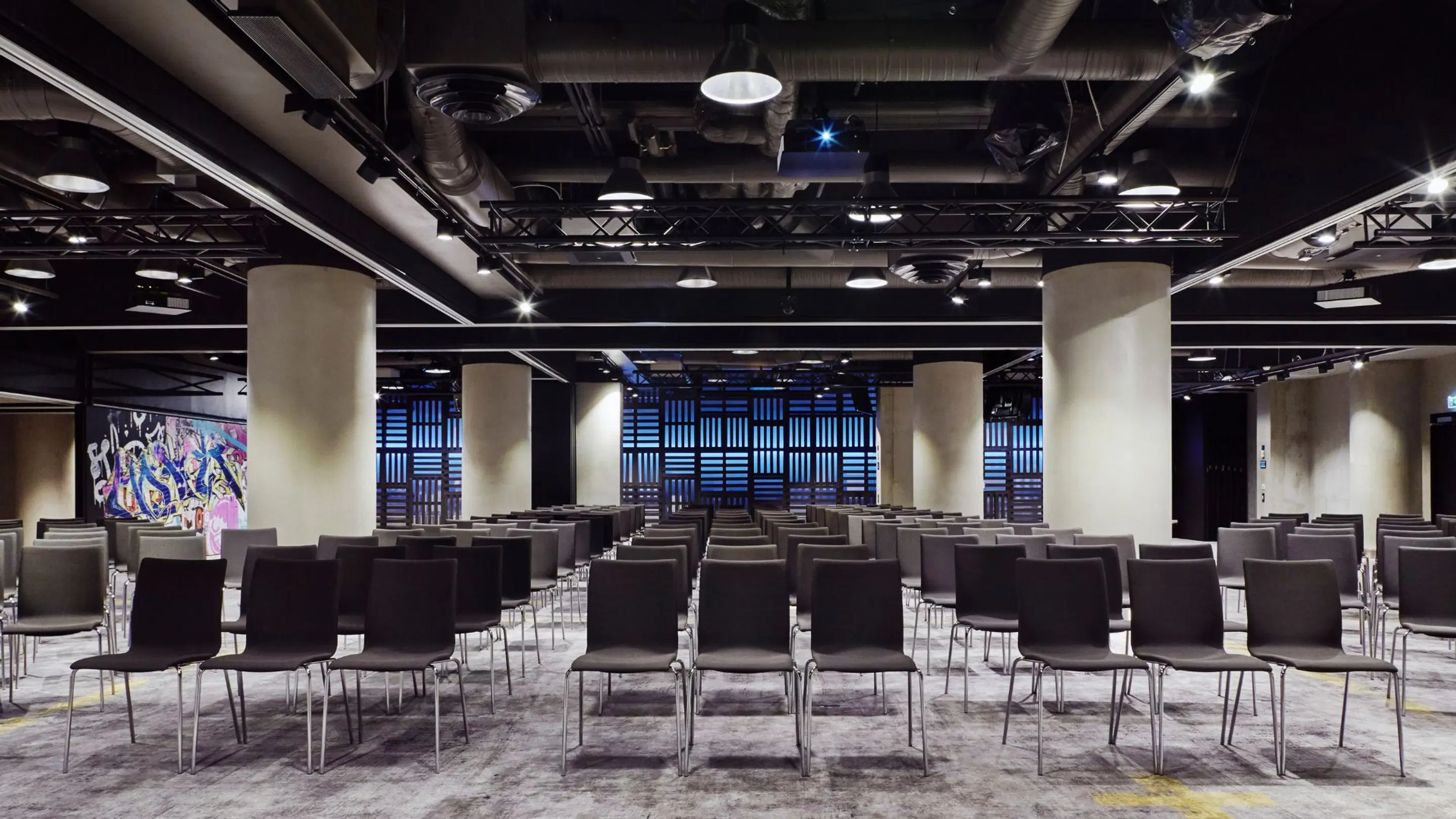 Modernt auditorium med stolar i rader, ljusa pelare och en blå bakgrundsvägg. Färgglad konst till vänster.