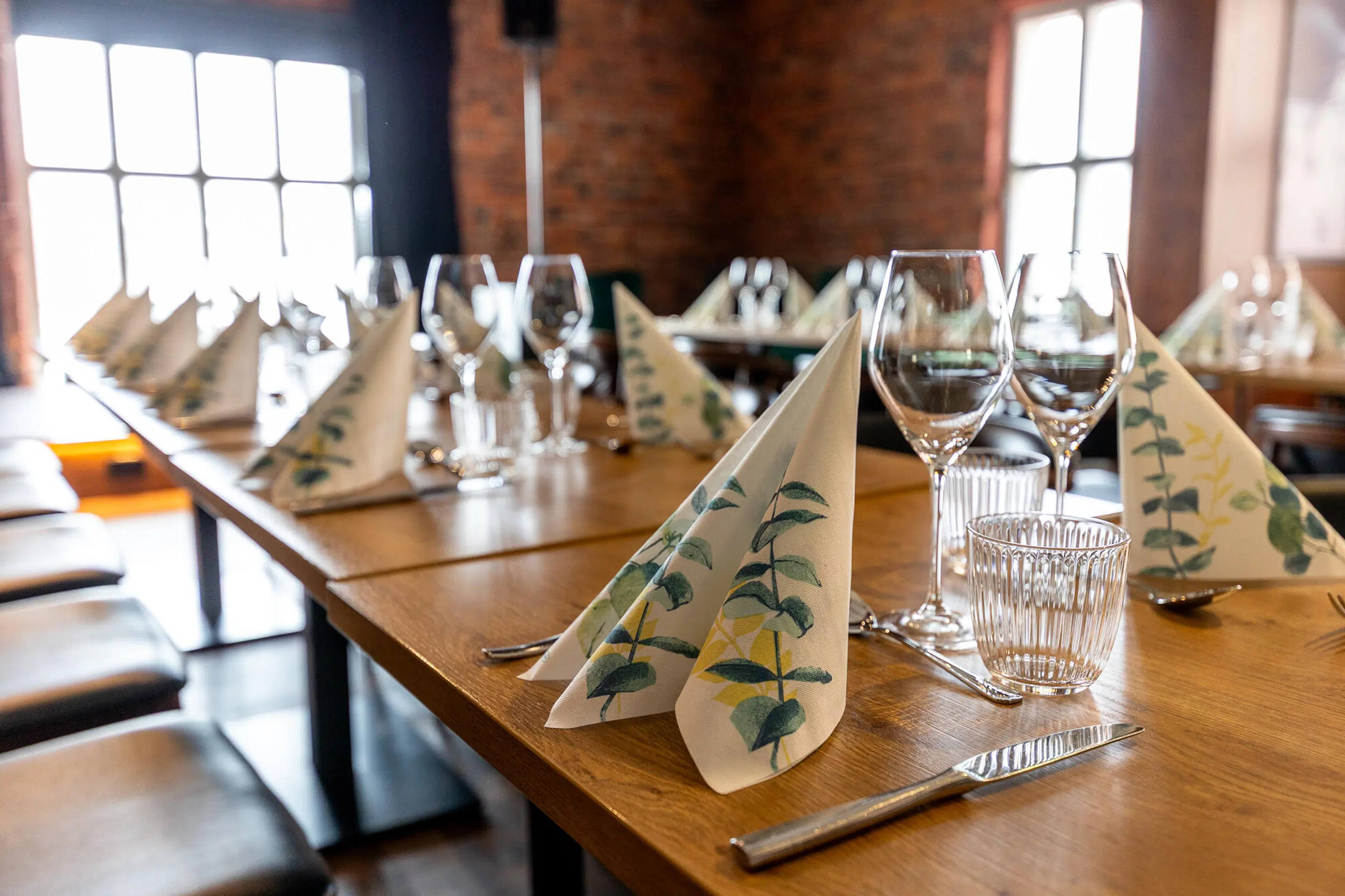 Restaurant tables set for dining. Wooden tables with plant-patterned napkins, wine glasses and hard flooring. Brick walls and large windows.