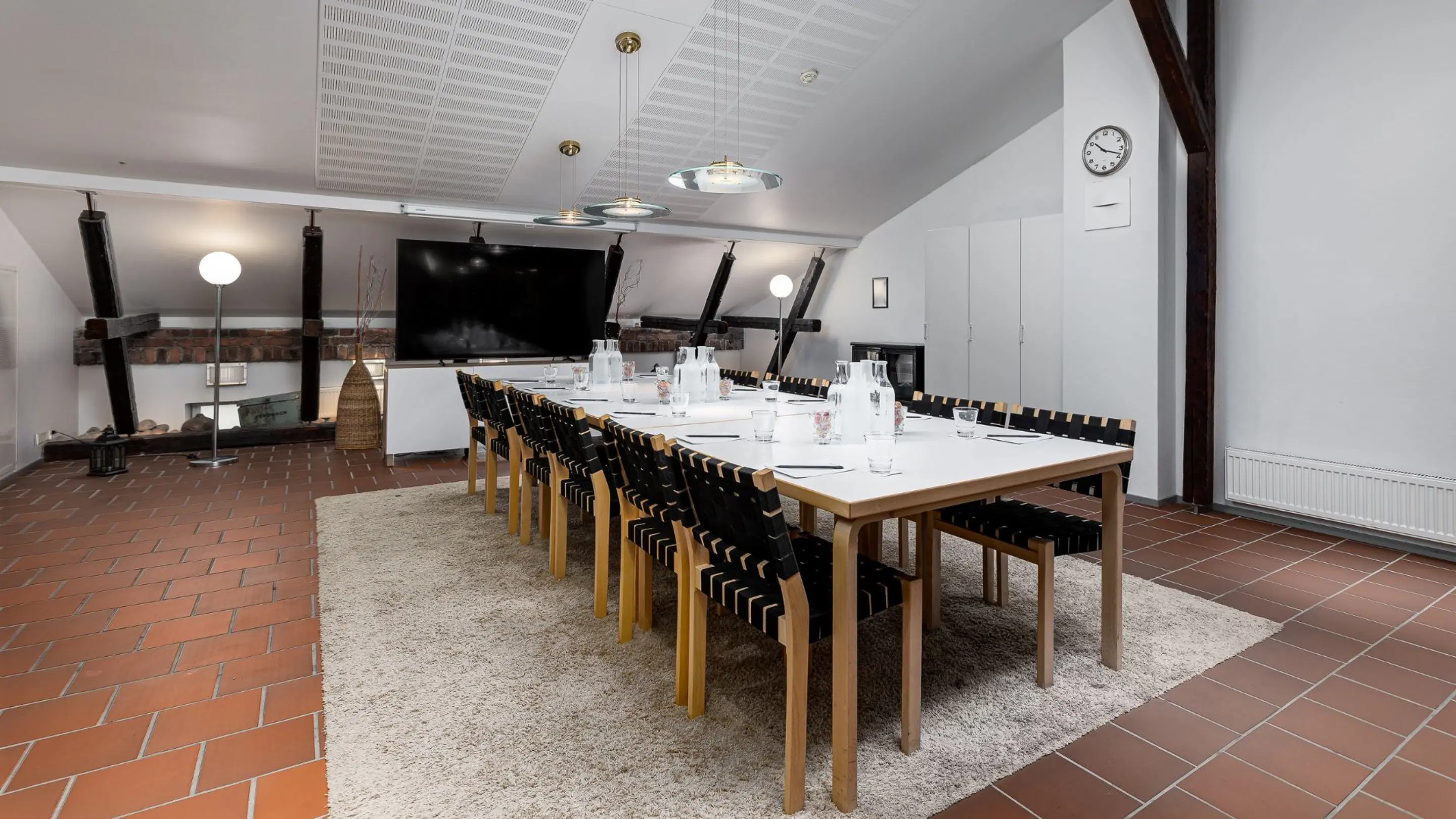 Modern meeting room with terracotta tile floor. Wooden table with black chairs on carpet, large screen in background. Exposed brick and wooden beams visible.