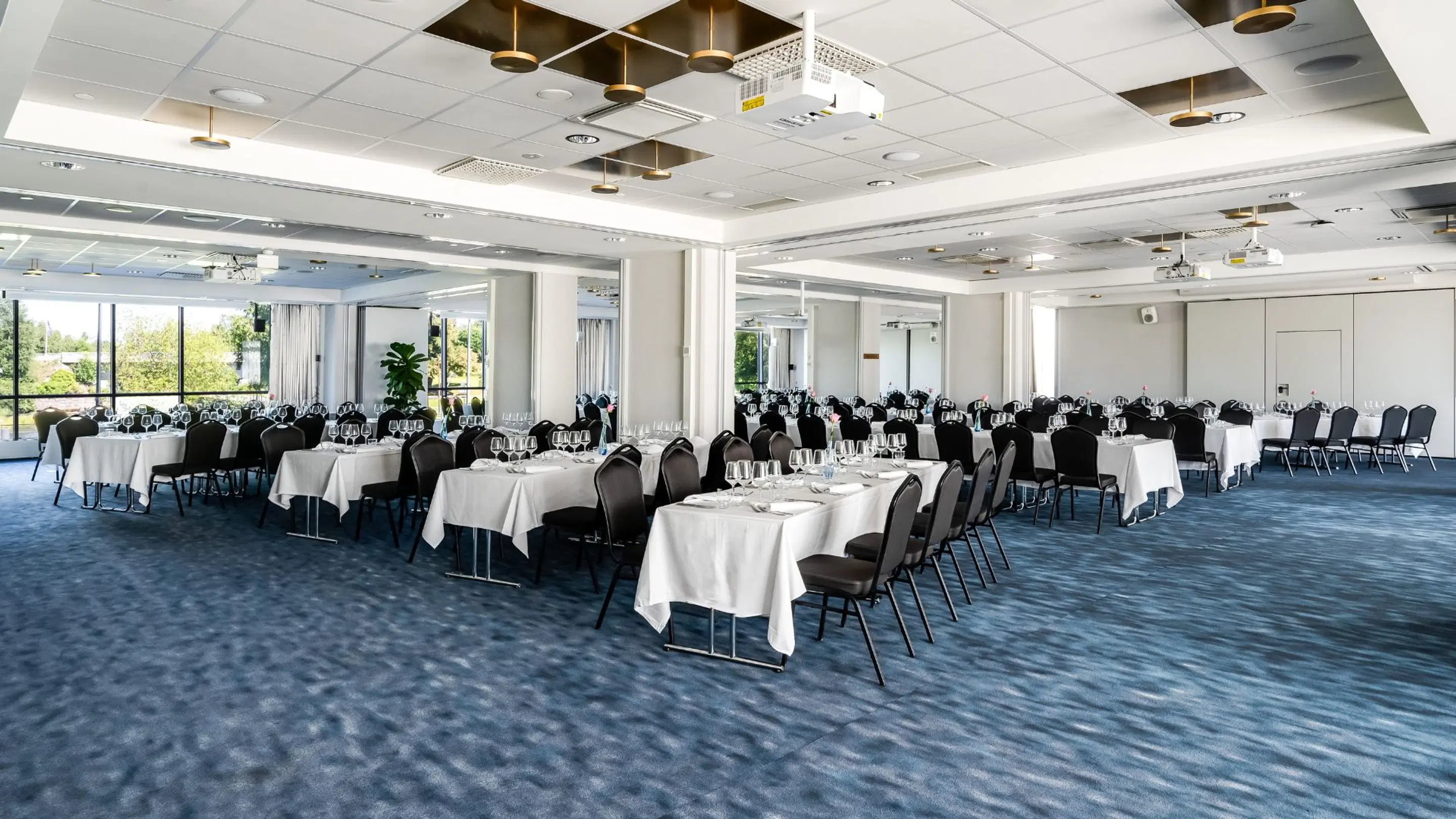 Spacious banquet hall with tables covered in white tablecloths and black chairs. Blue carpeted floor, large windows, and ceiling-mounted projector.
