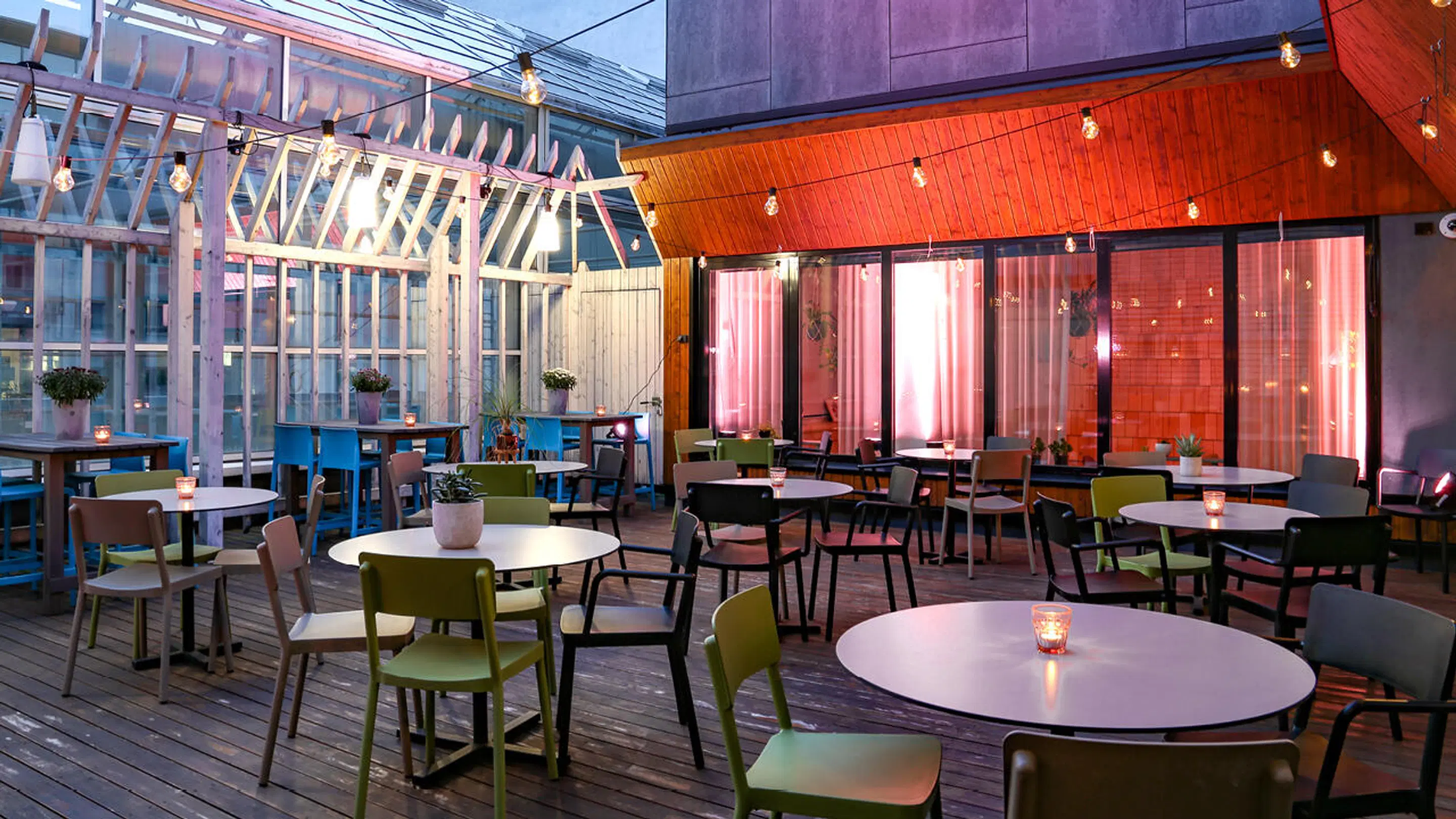 Restaurangterrass med trägolv, runda bord och stolar. Glastak och väggar, dekorativ belysning och gröna växter. Modern inredning med träpaneler.