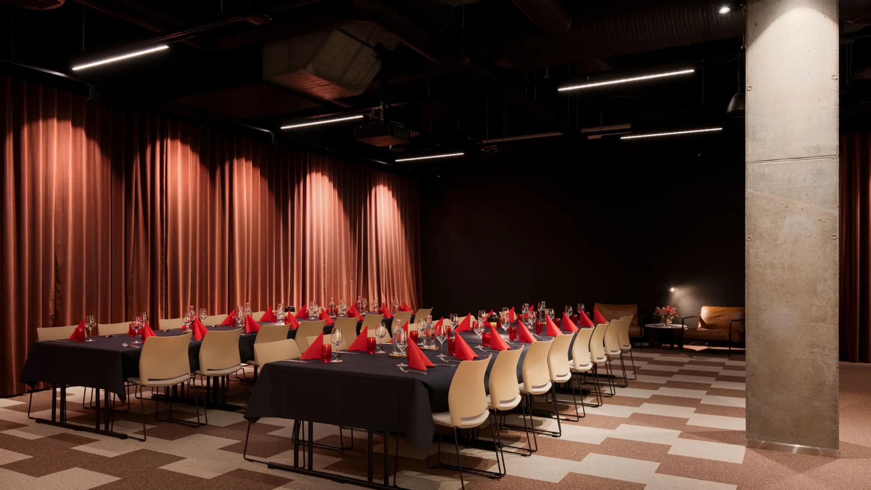 Dining area set up for an event with long tables covered in black tablecloths, light-colored chairs, and red napkins. Red curtains in background, checkered floor.