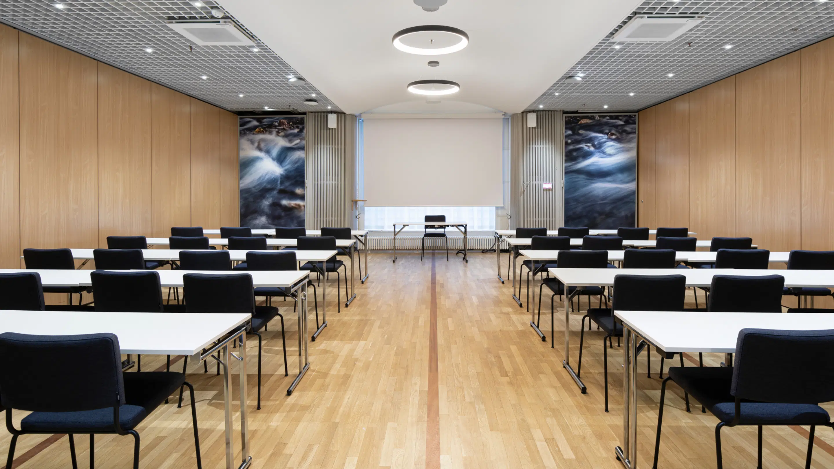 Modernt konferensrum med träpanelväggar, ljust trägolv, vita bord och svarta stolar, projektorduk framme och konstverk föreställande flödande vatten på väggarna.
