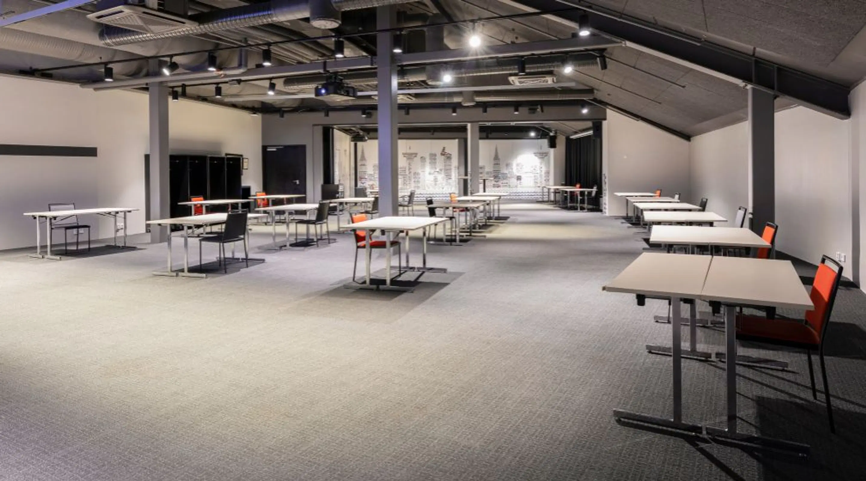 Avara konferenssi- tai työtila, jossa on tasainen mattolattiapinta, matala katto näkyvillä putkilla ja pilarein jäsennelty. Tilassa on hajallaan työpöytiä ja tuoleja.