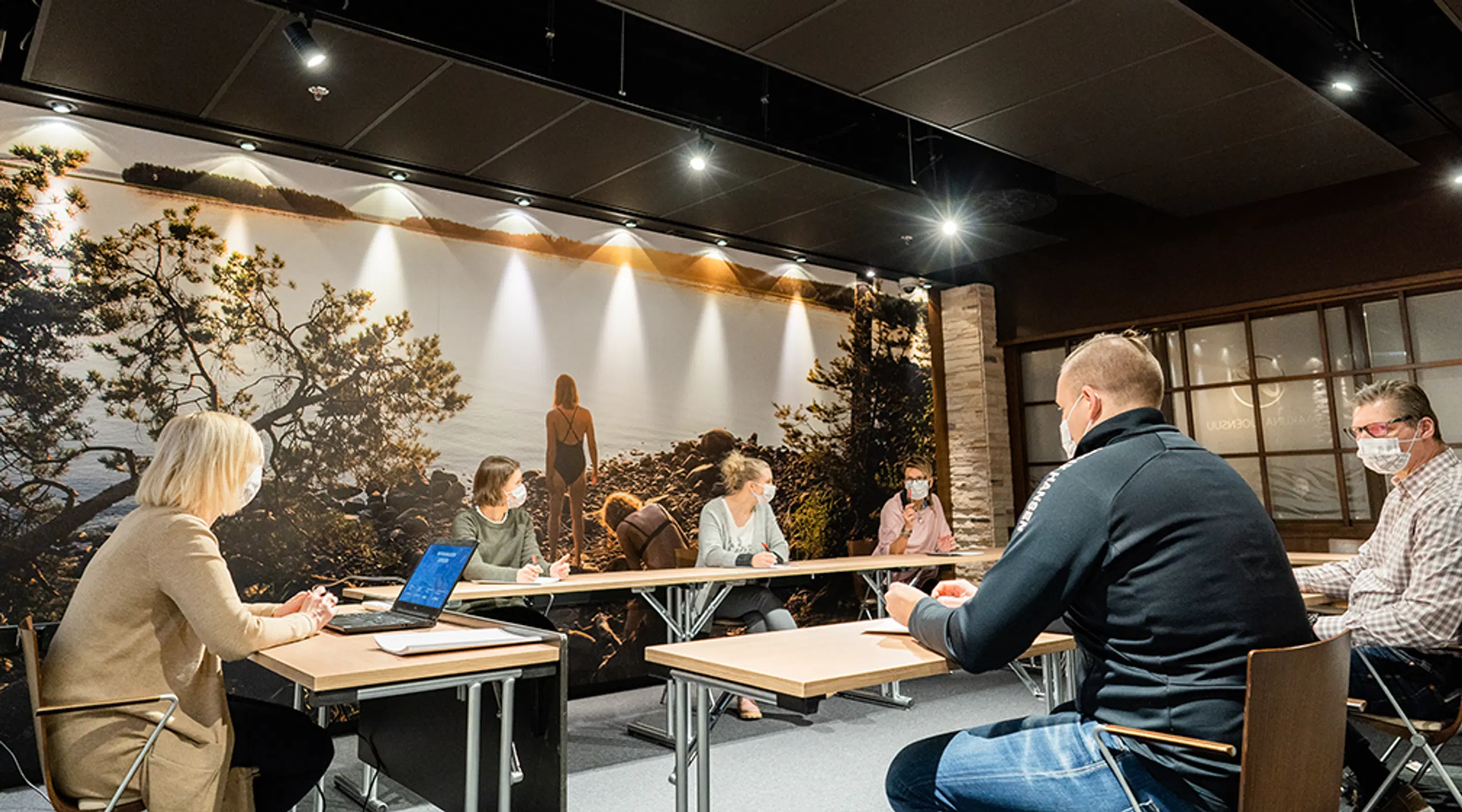 Meeting room with people seated around a table, large forest landscape mural on the wall behind them. Some wearing face masks.