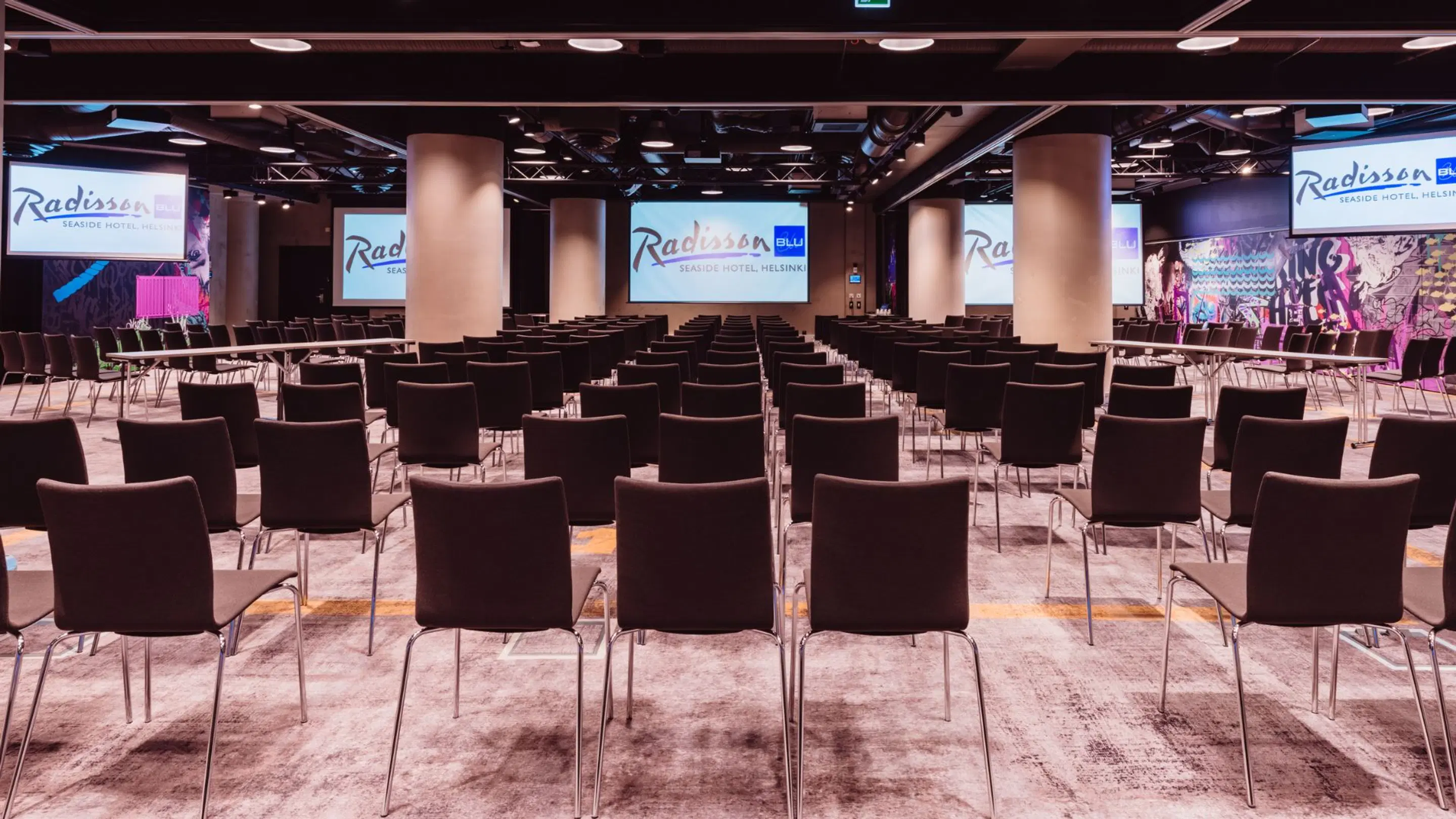 Rymlig konferensauditorium i Radisson-hotellet med svarta stolar i rader, stora skärmar framme och höga pelare. Plant golv, bra belysning.