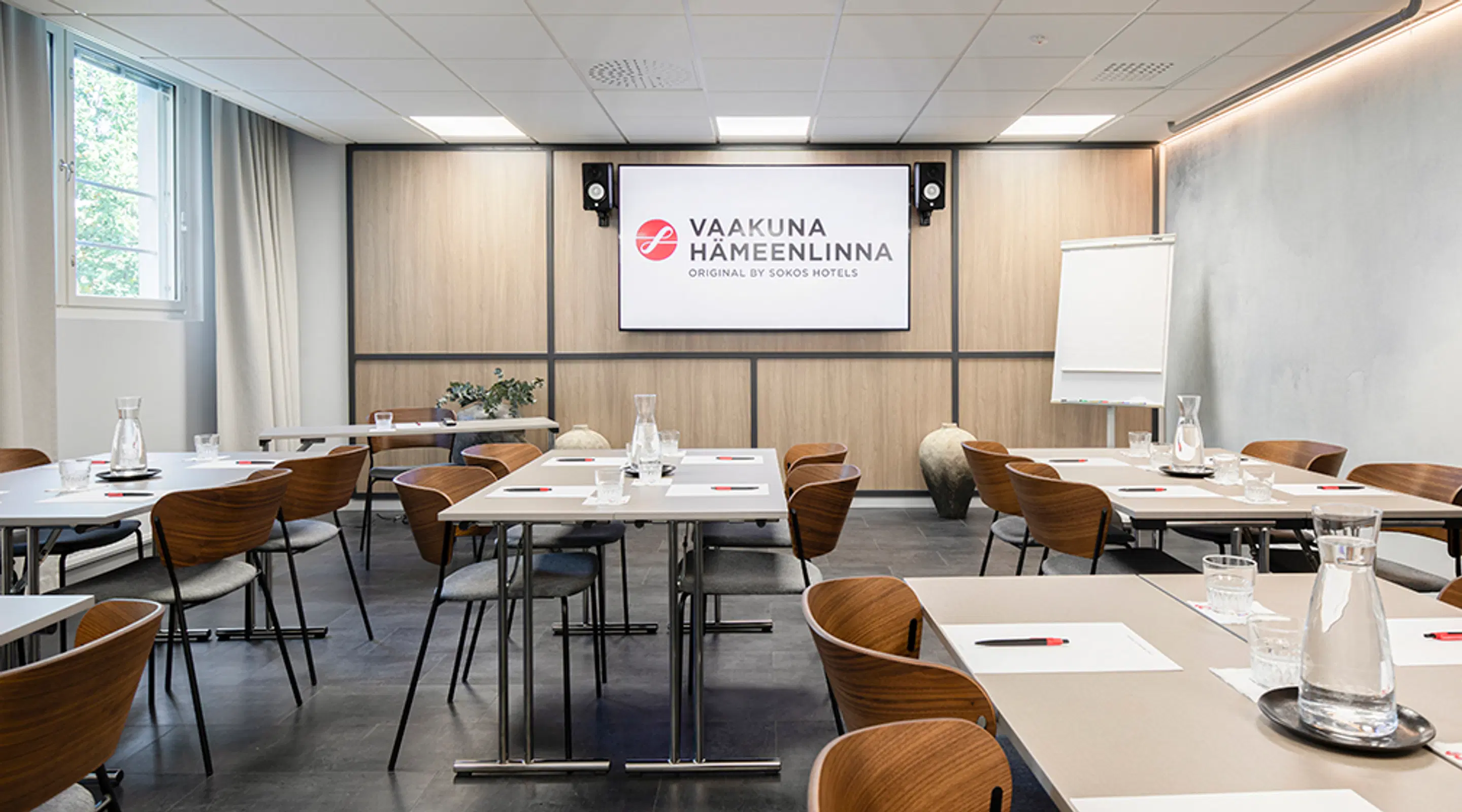 Ljust konferensrum med träbord och vita bord. Presentationsskärm framme, grönska syns genom fönstret, blädderblock på sidan.