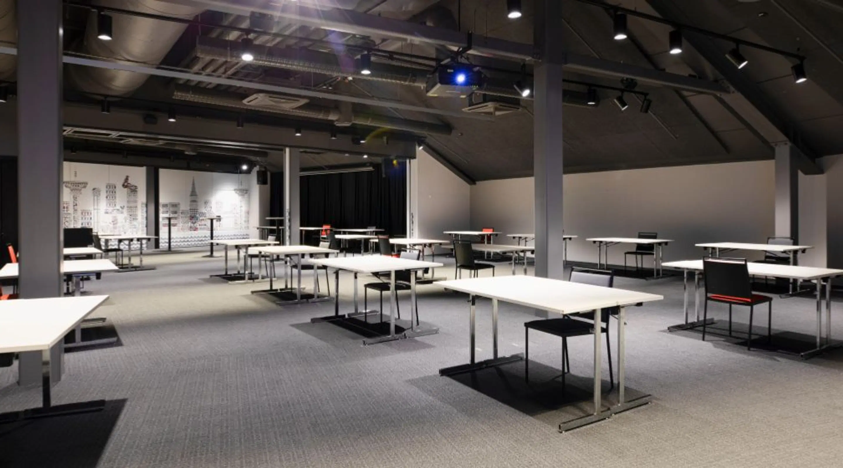 Moderni kokoustila, jossa pitkiä valkoisia pöytiä ja mustia tuoleja lyhytpinnoitteisella harmaalla moquette-lattialla. Korkea katto teollisella ilmastointikanavoinnilla ja hyvä valaistus.