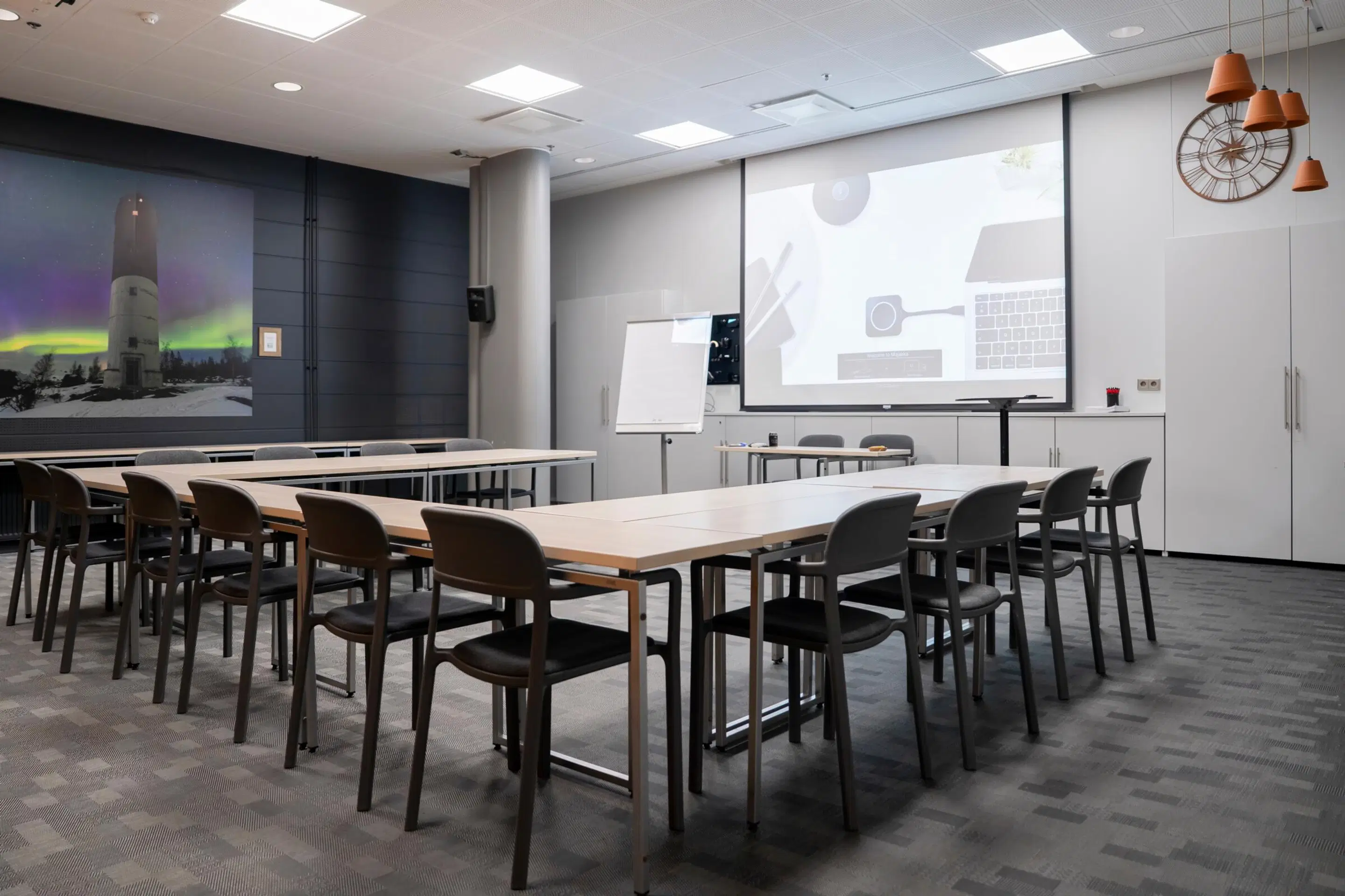 Modern meeting room with wooden tables arranged in rows, chairs, whiteboard, and projector screen. Aurora image on wall, patterned carpeted floor.