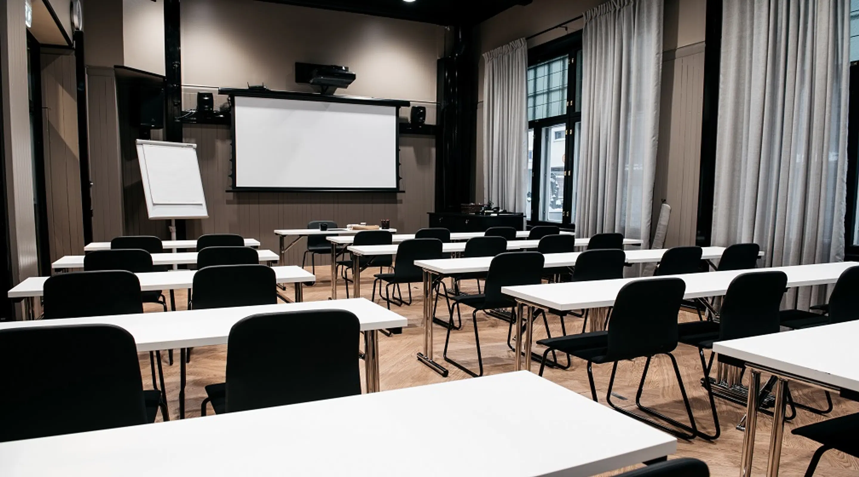 Conference room with white tables and black chairs in rows, projection screen at front, whiteboard, and windows covered with light curtains.