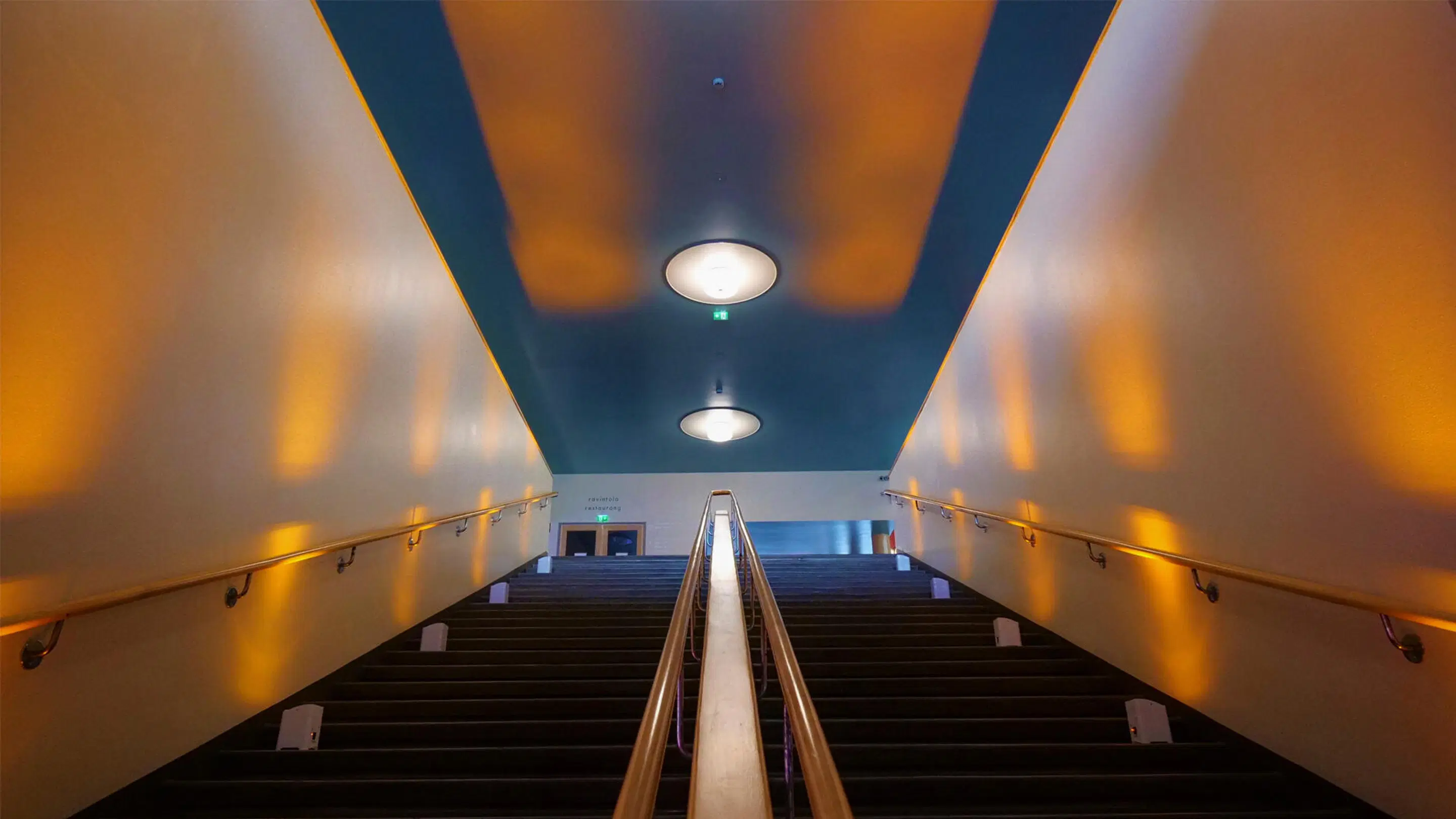 Wide staircase with a metal center railing and handrails along the sides. The ceiling is blue and walls have warm yellow lighting.