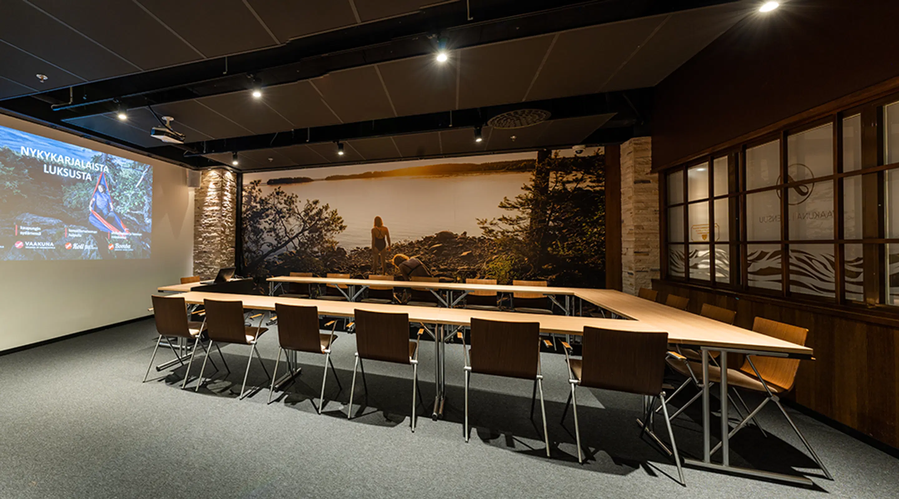 Modern meeting room with U-shaped table arrangement and chairs, nature photograph on wall, projector screen, good lighting and level flooring.