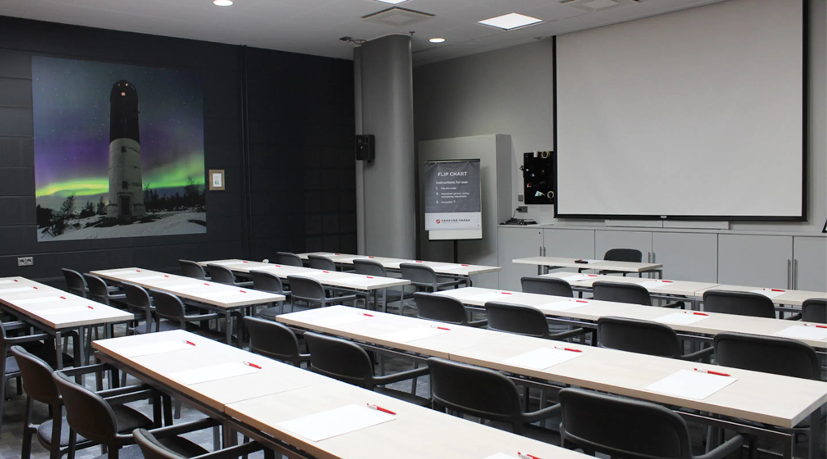 Classroom with long tables and chairs. Large image of a lighthouse under northern lights on the wall, projection screen at the front.