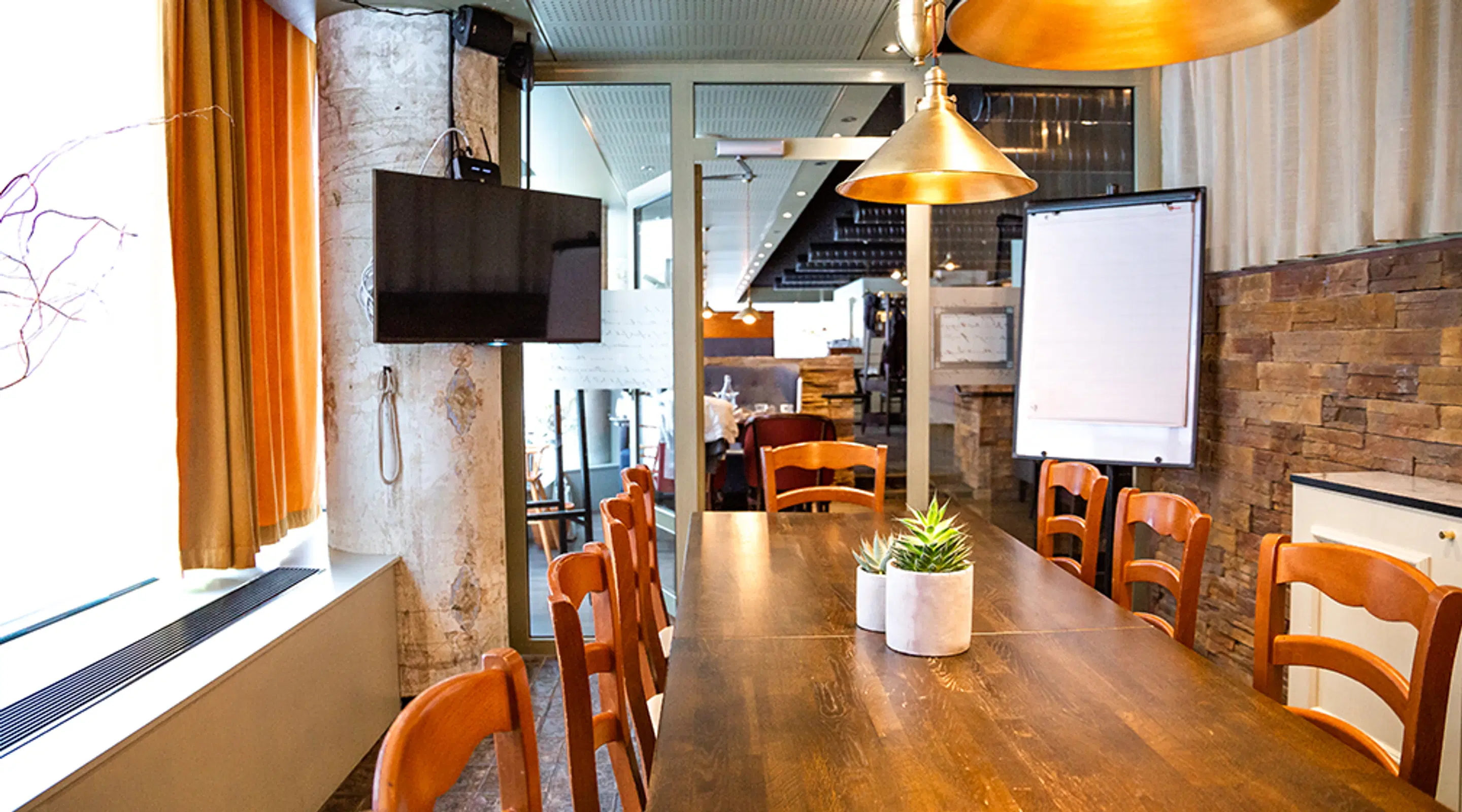 Meeting room with wooden table, orange chairs and hanging gold lamps. The room has a whiteboard, TV, and brick walls.