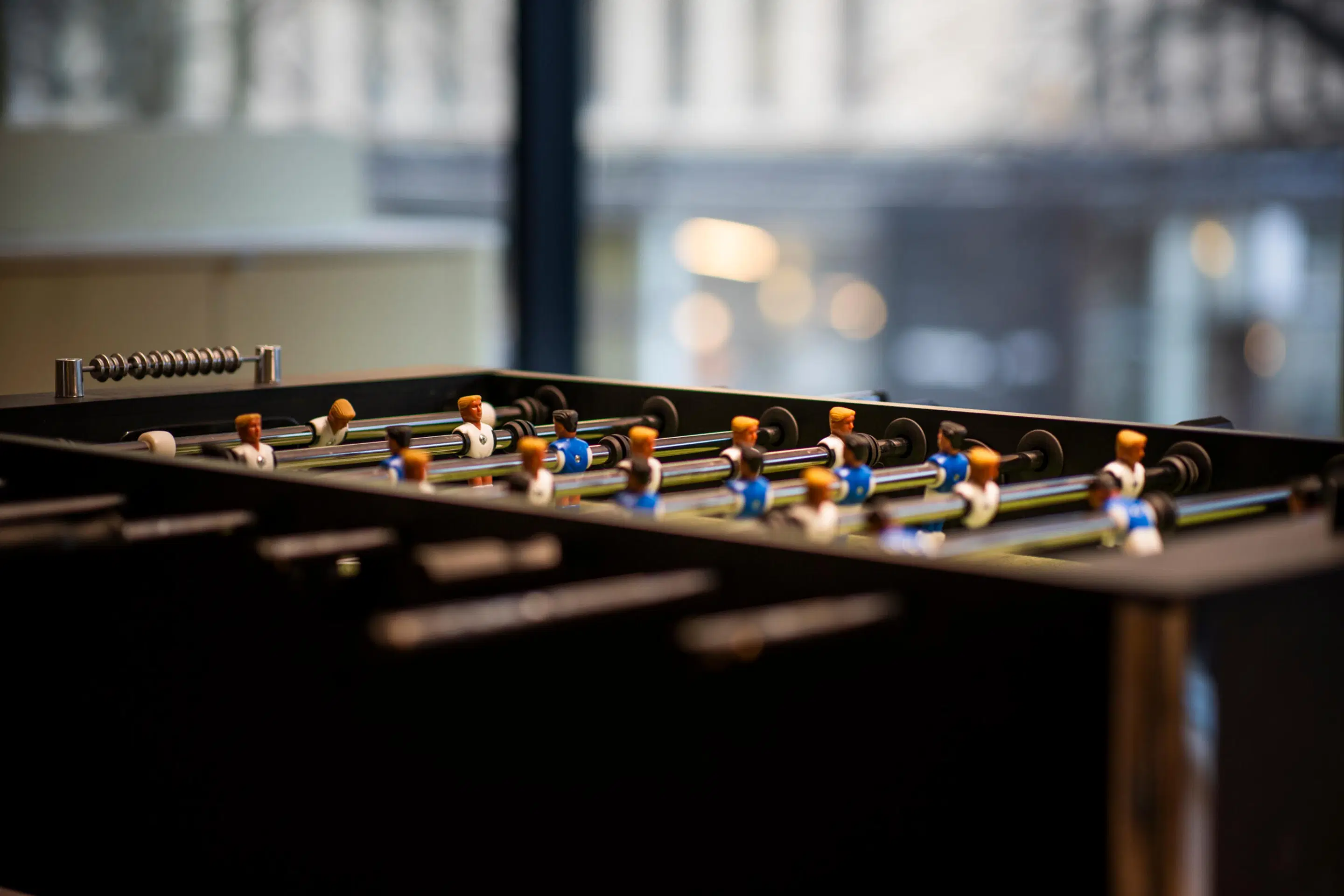 Foosball table with blue and white player figures mounted on rods on a black table. Blurry window view in background.