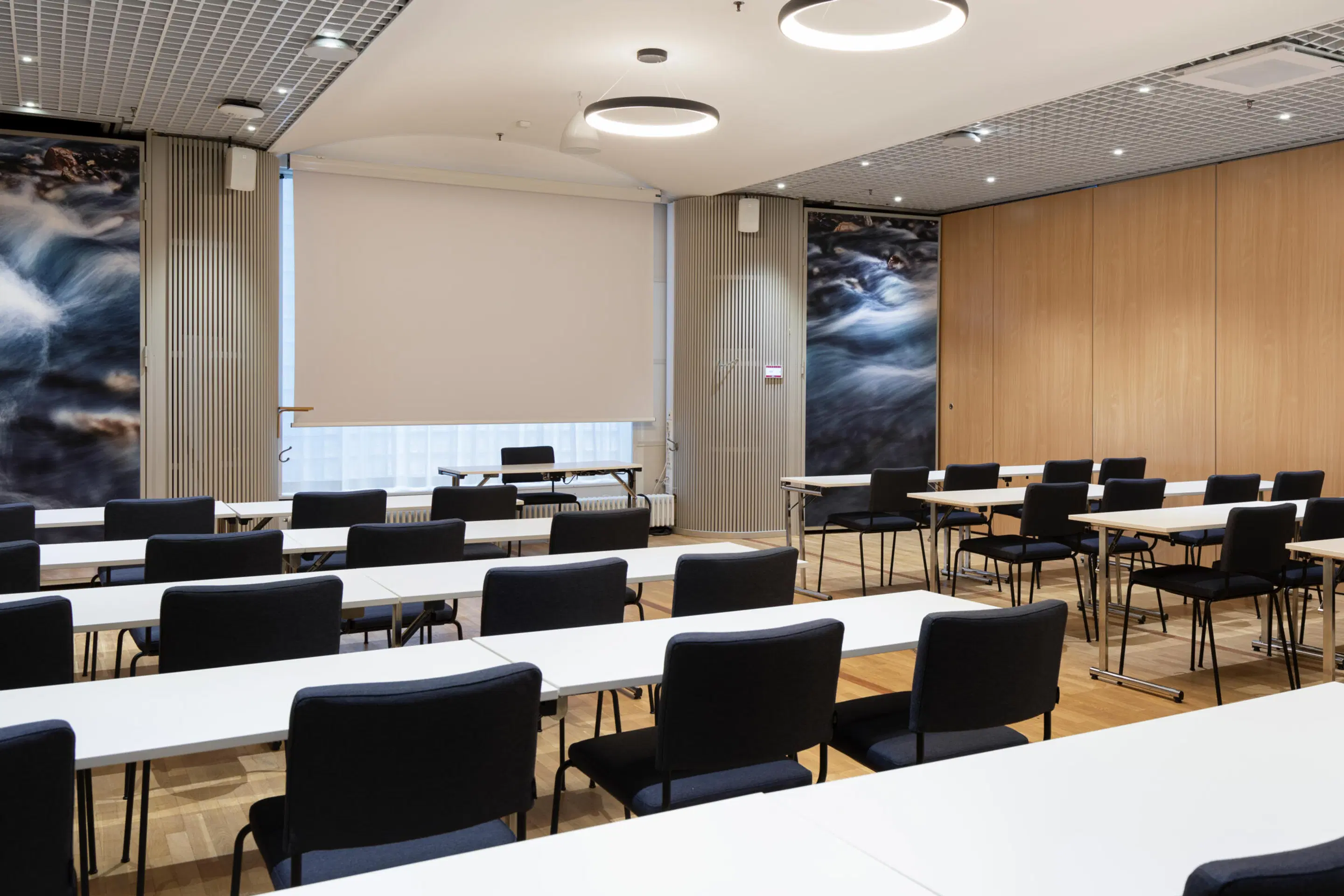 Modern meeting room with white tables and black chairs. Flowing water images on walls, wooden floor and circular ceiling lights.