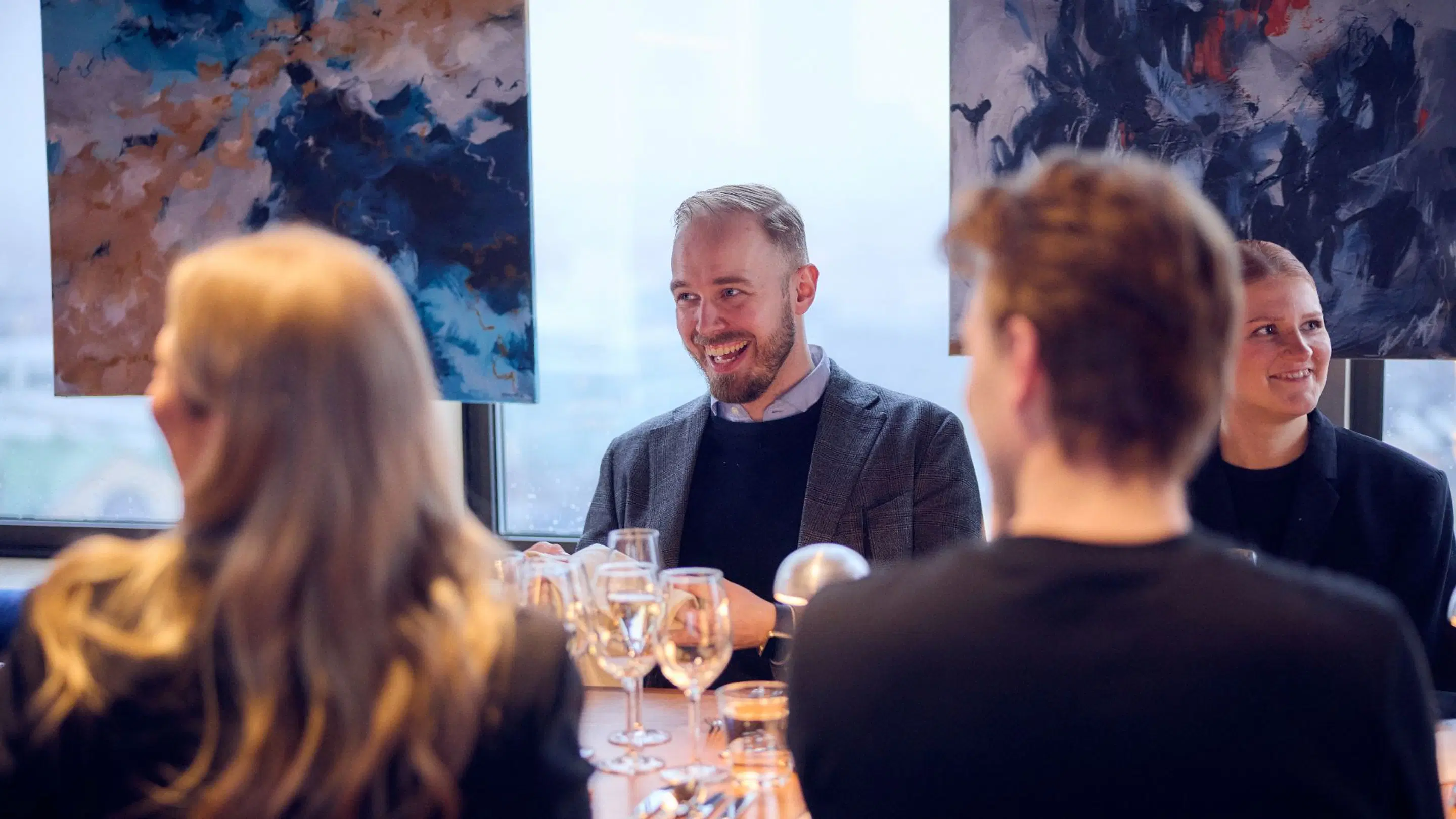 Personer njuter av drycker vid ett bord med färgglada abstrakta konstverk och fönsterutsikt i bakgrunden.