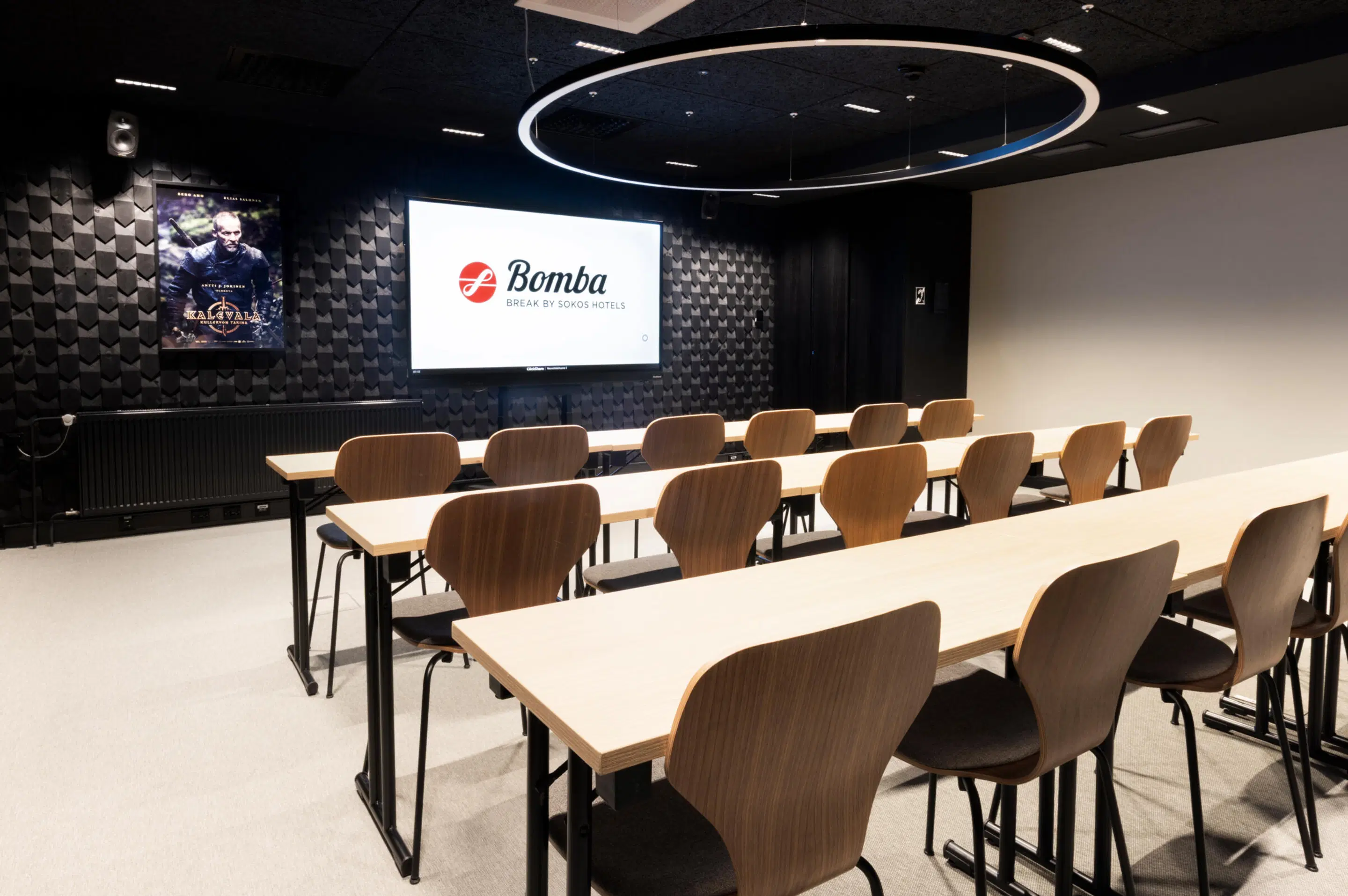 Modern conference room with rows of tables and chairs, screen at front, black patterned wall and level floor surface.