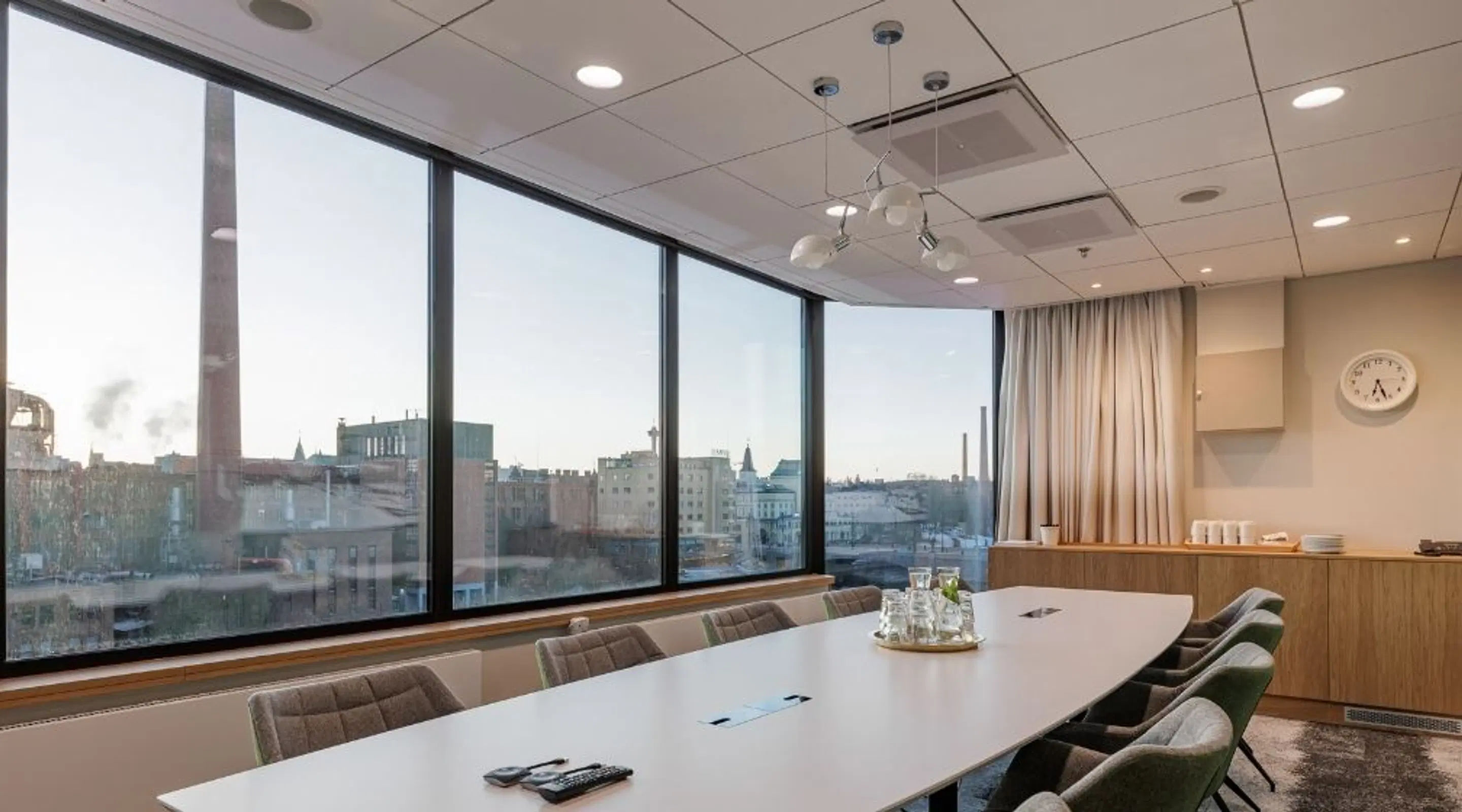 Modern meeting room with large window wall overlooking city view. Light-colored table, gray chairs and a serving station.