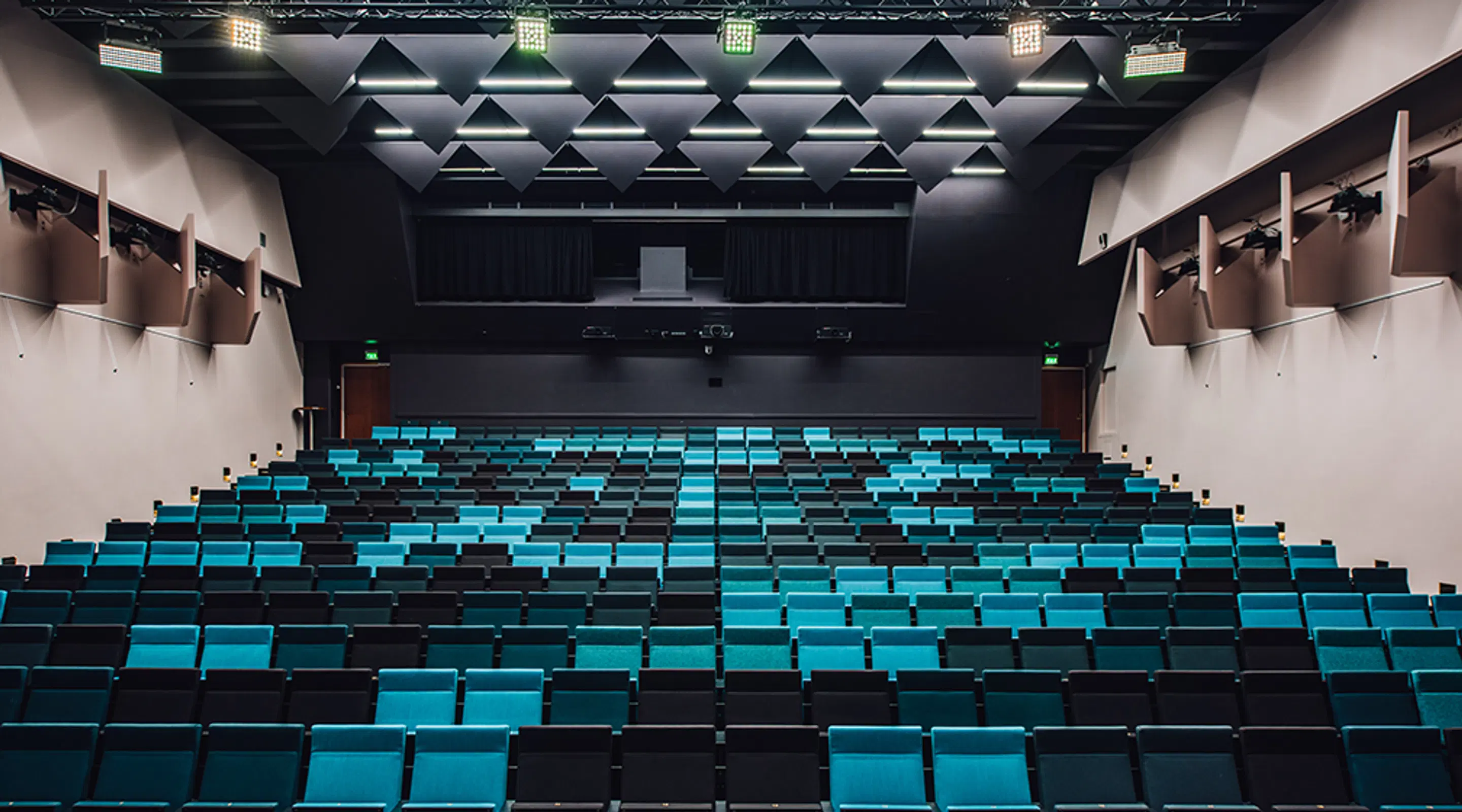 Moderni auditorio, jossa tyylikkäitä tealinsinisiä ja mustia istuinrivejä, keskikäytävä portaineen, korkealla kattotrussilla varustettu valaistus ja näyttämö taustalla.