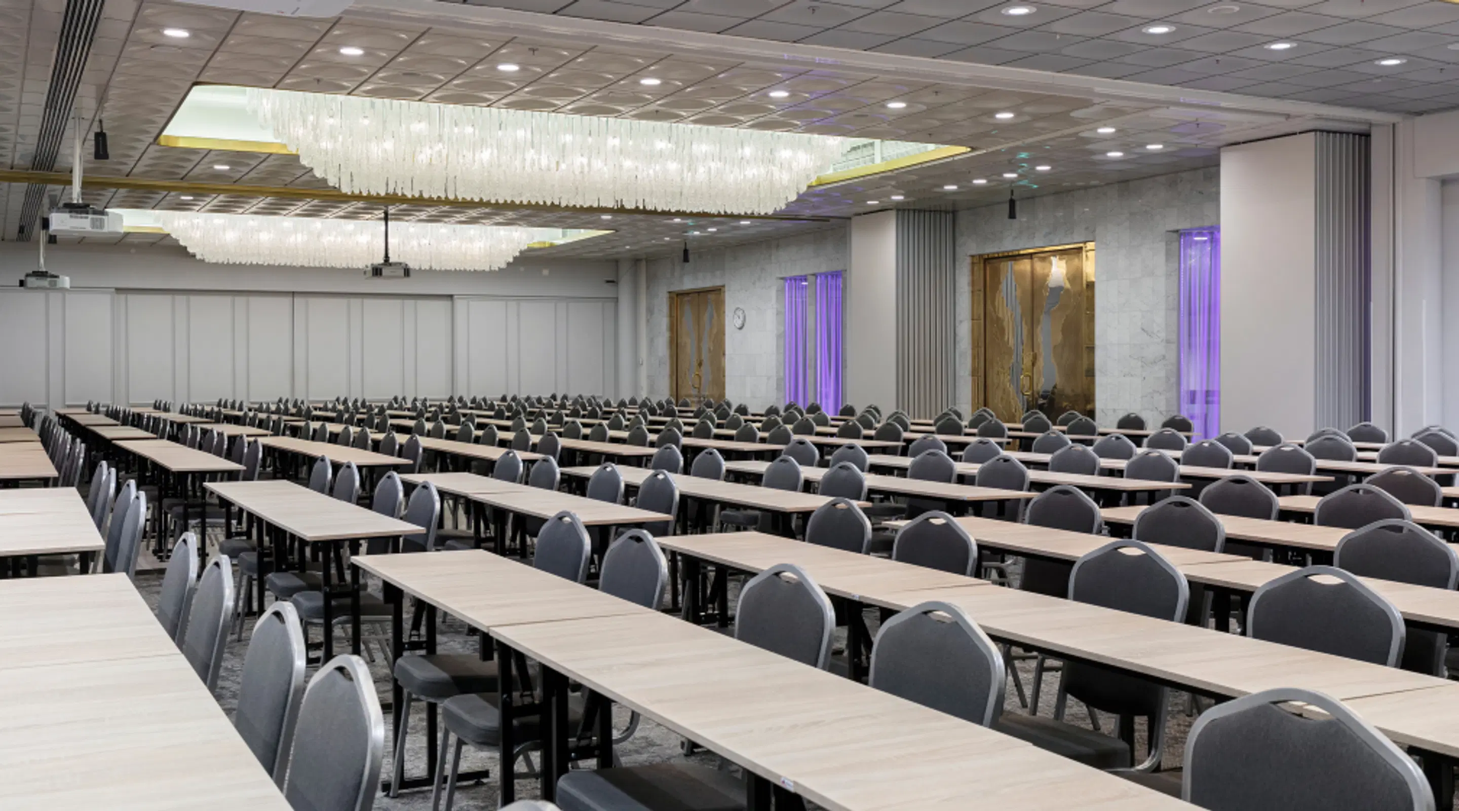 Large conference room with long tables and grey chairs arranged in rows. Decorative ceiling lights and purple lighting along walls.