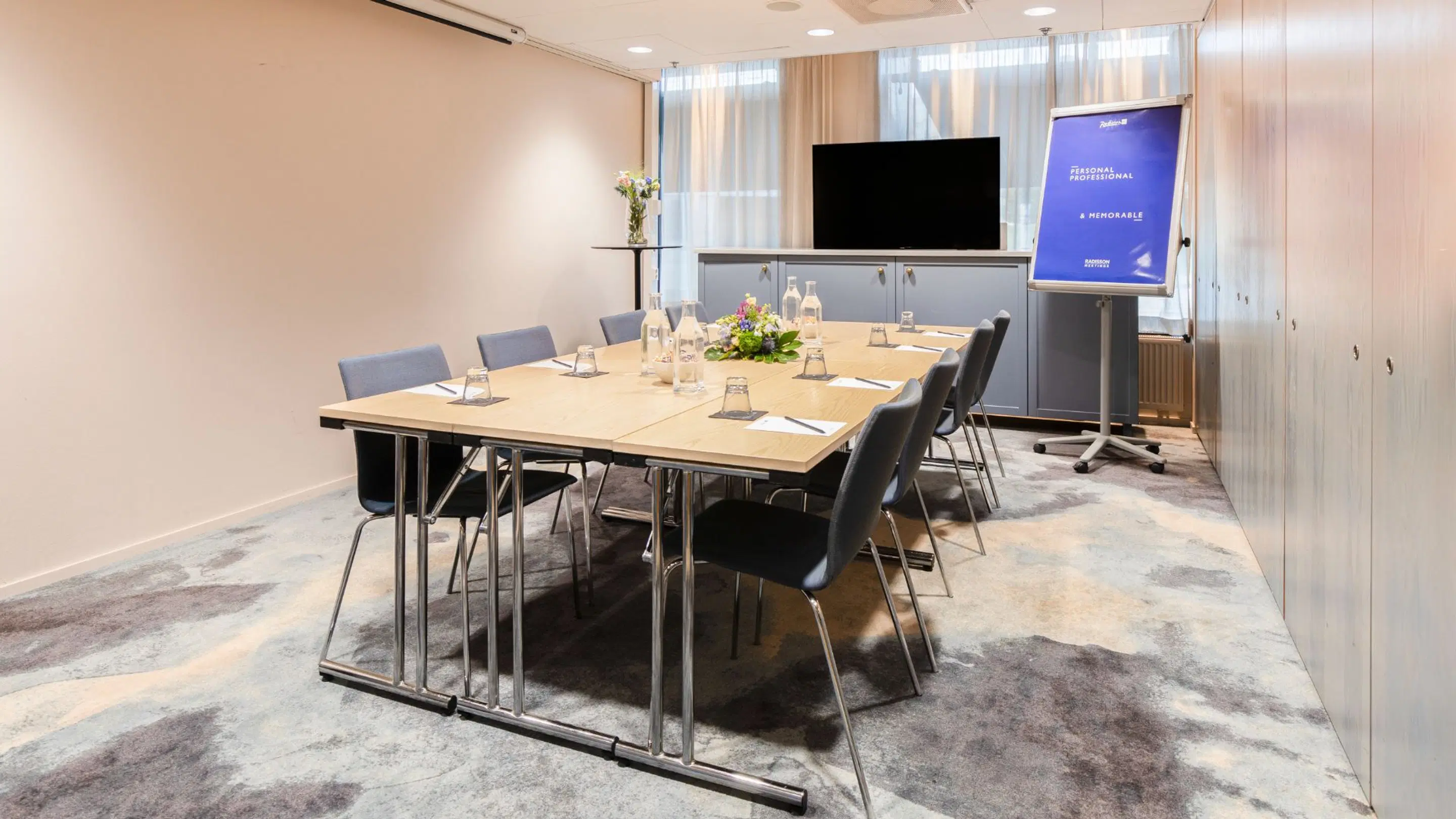 Modernt konferensrum med träbord, blå stolar, tv och presentationsskärm. Rummet har heltäckningsmatta och god belysning.