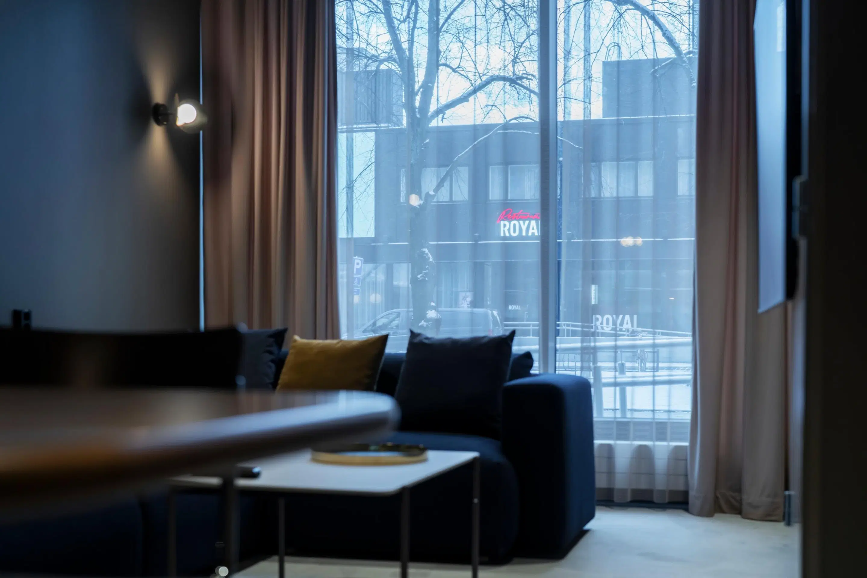 Hotellituba sinise diivaniga, millel on kollane padi, suured aknad ja vaade teisele hoonele. Toas on tasane põrand ja hea valgustus.