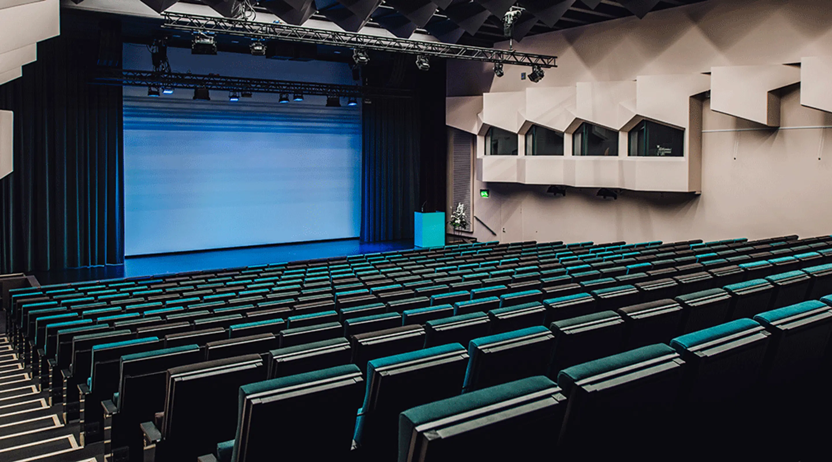 Moderni auditorio, jossa on turkoosit istuimet, suuri valkokangas ja sinisellä valolla valaistu näyttämö.