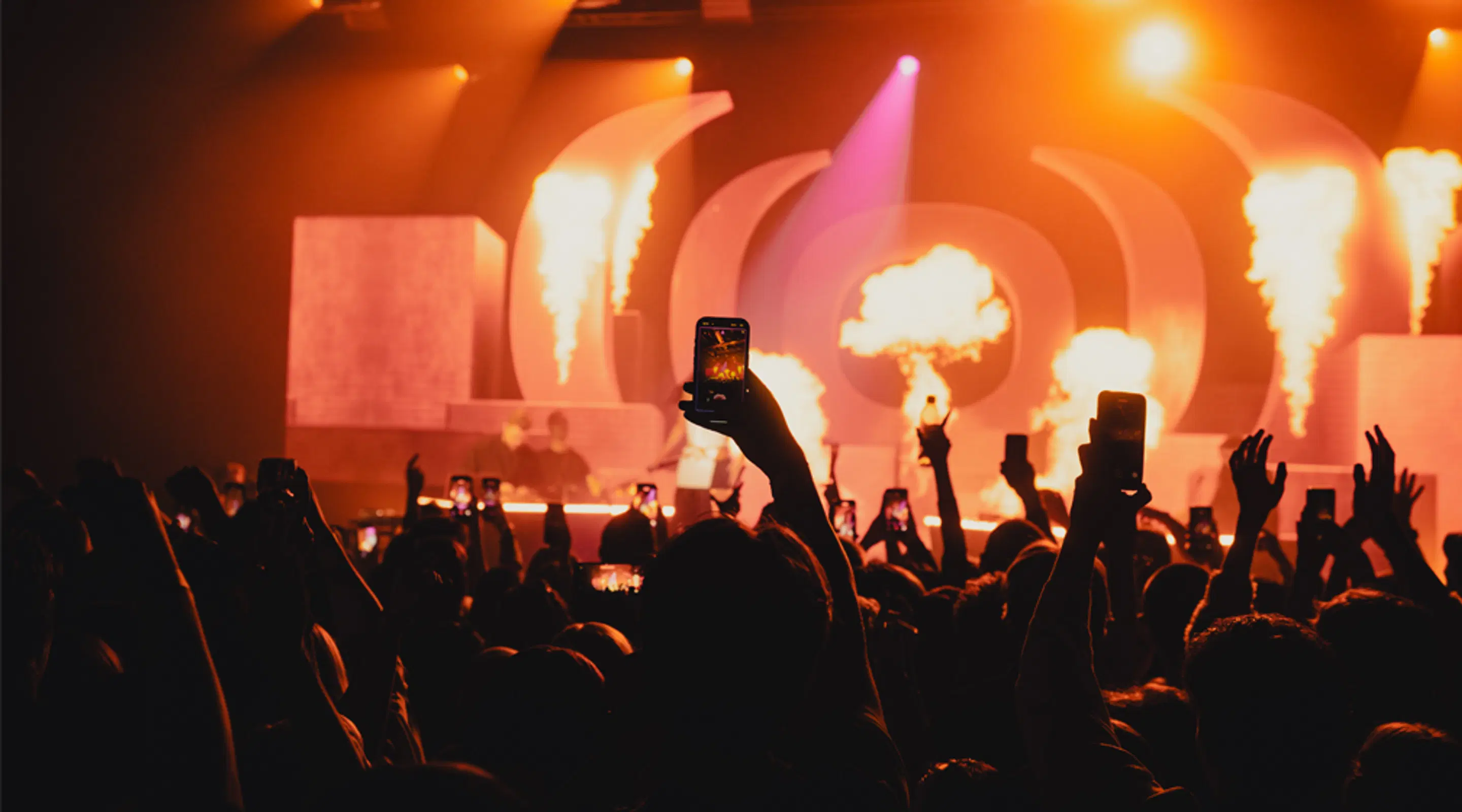 Concert crowd holding phones in the air while fire effects and colorful lights illuminate the stage in orange lighting.