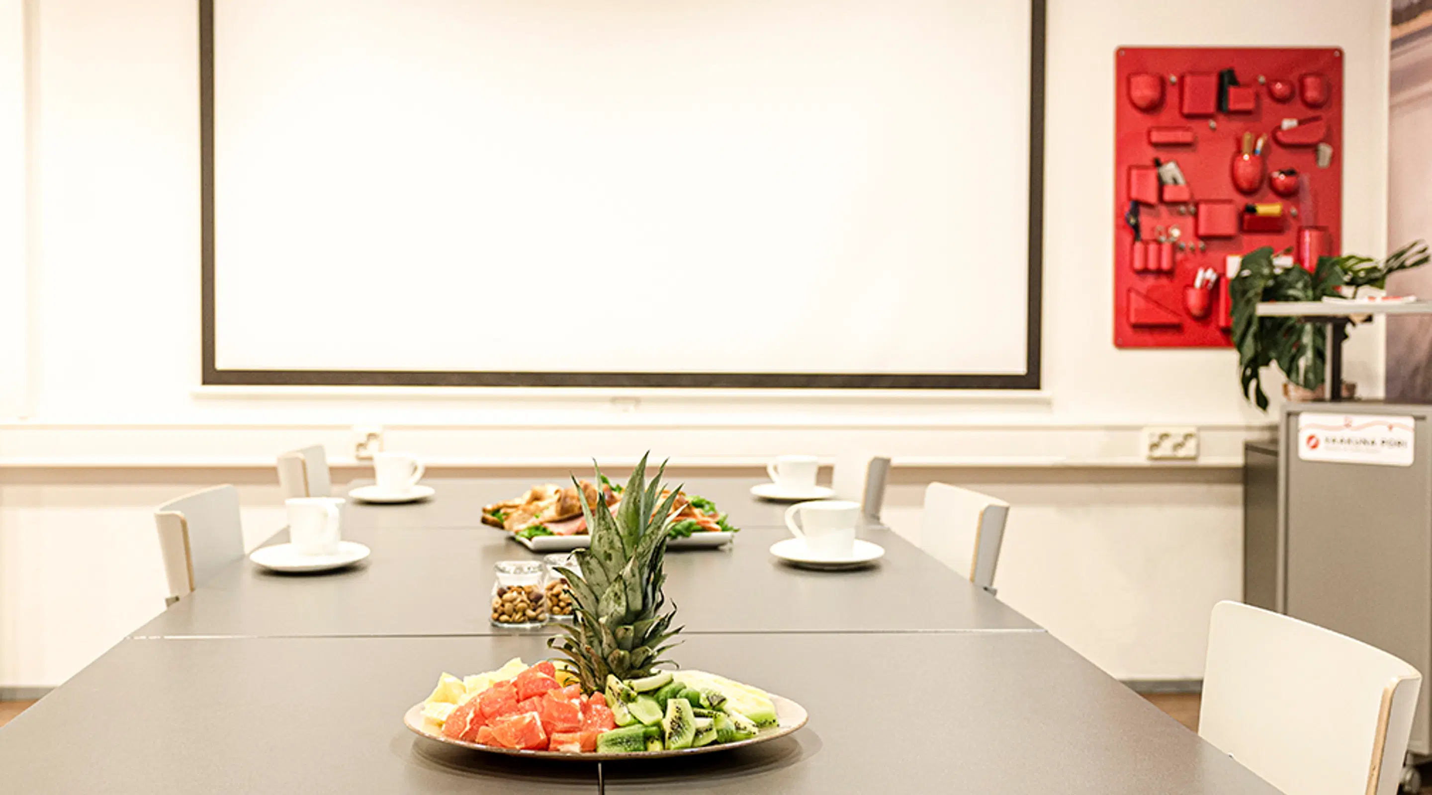 Conference table with fruit platter featuring pineapple and watermelon. White projection screen and red bulletin board in background.