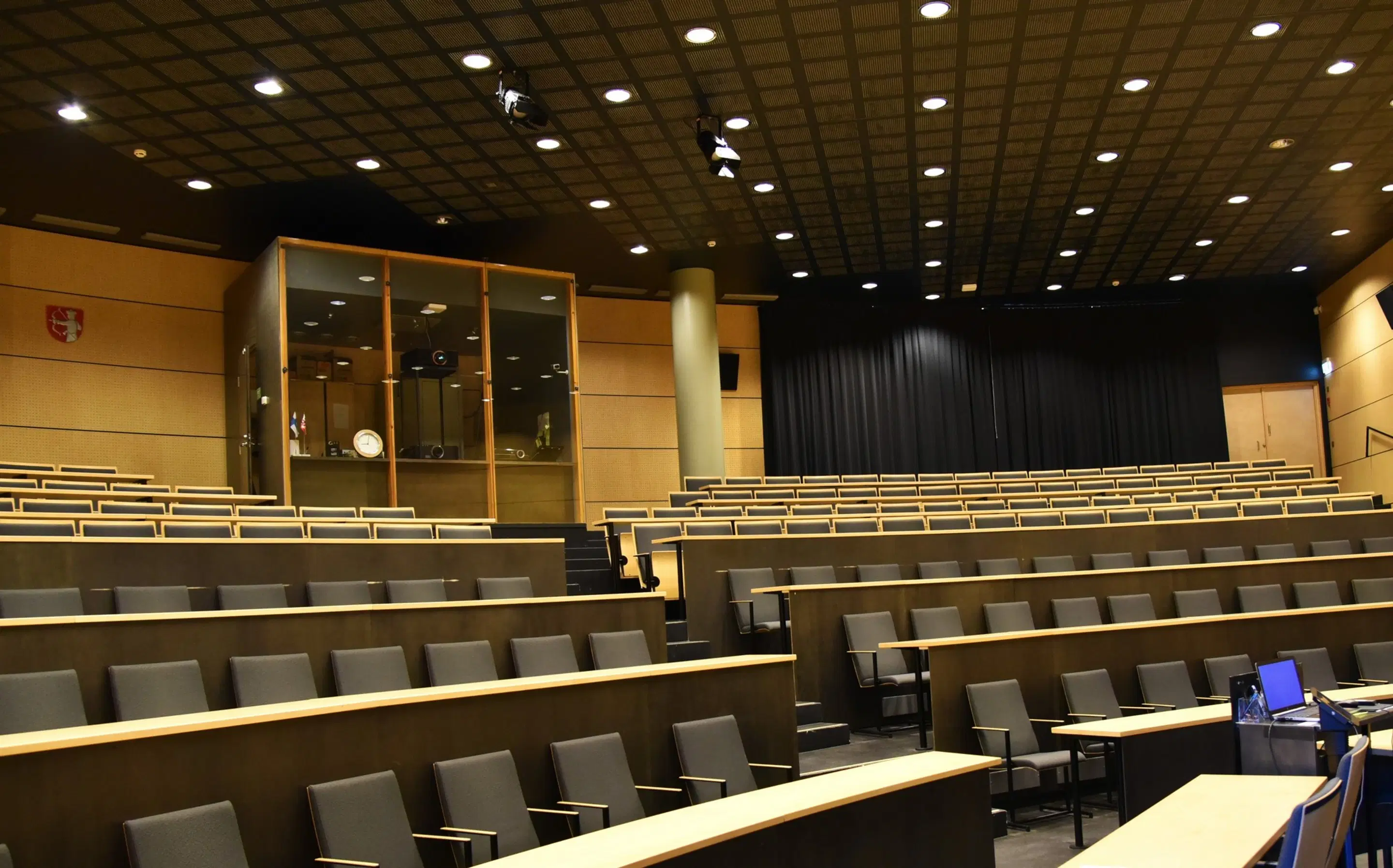 Auditoorium astmeliste istumisridade, laua-pindade ja hallide toolidega. Ridade vahel on trepid. Laes on prožektorid ja taga must kardin.