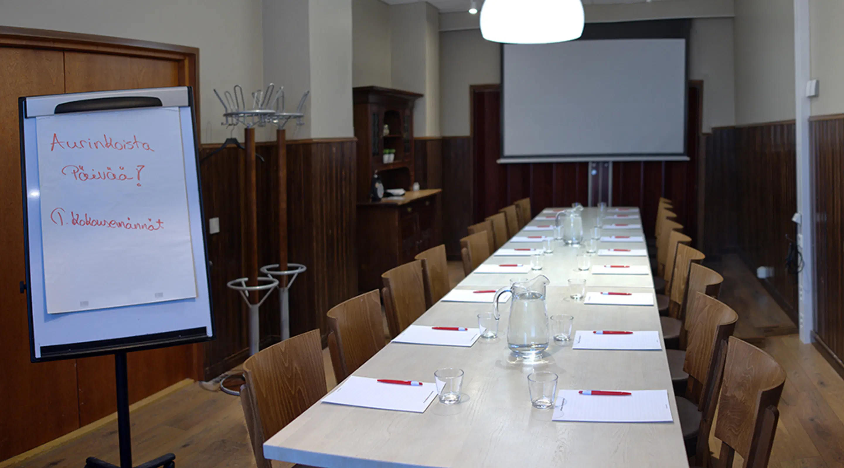 Meeting room with wooden chairs and long table set with notepads and water pitchers. Projection screen visible in background.
