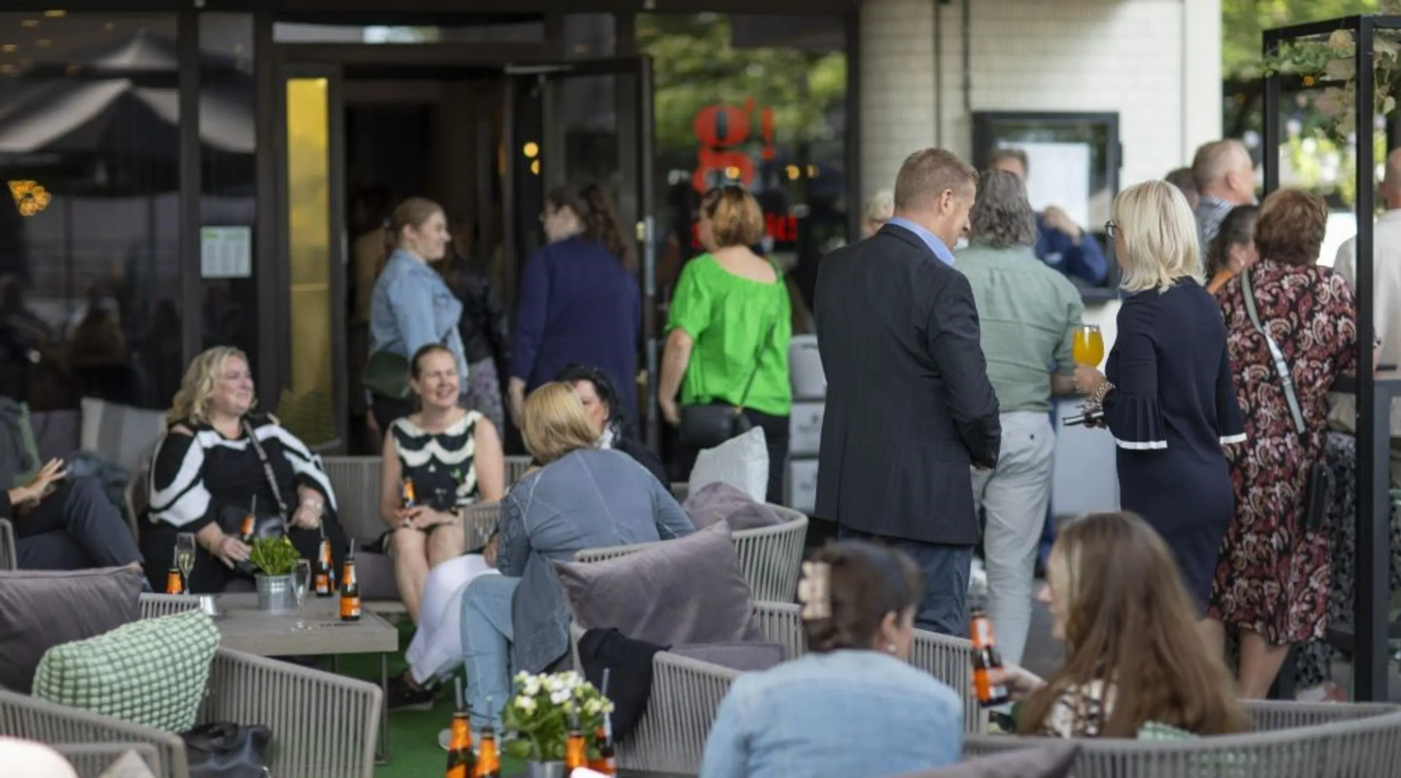 Personer på en trevlig terrass eller restaurangområde med ljusa rottingstolar där folk sitter och njuter av drycker och samtal.