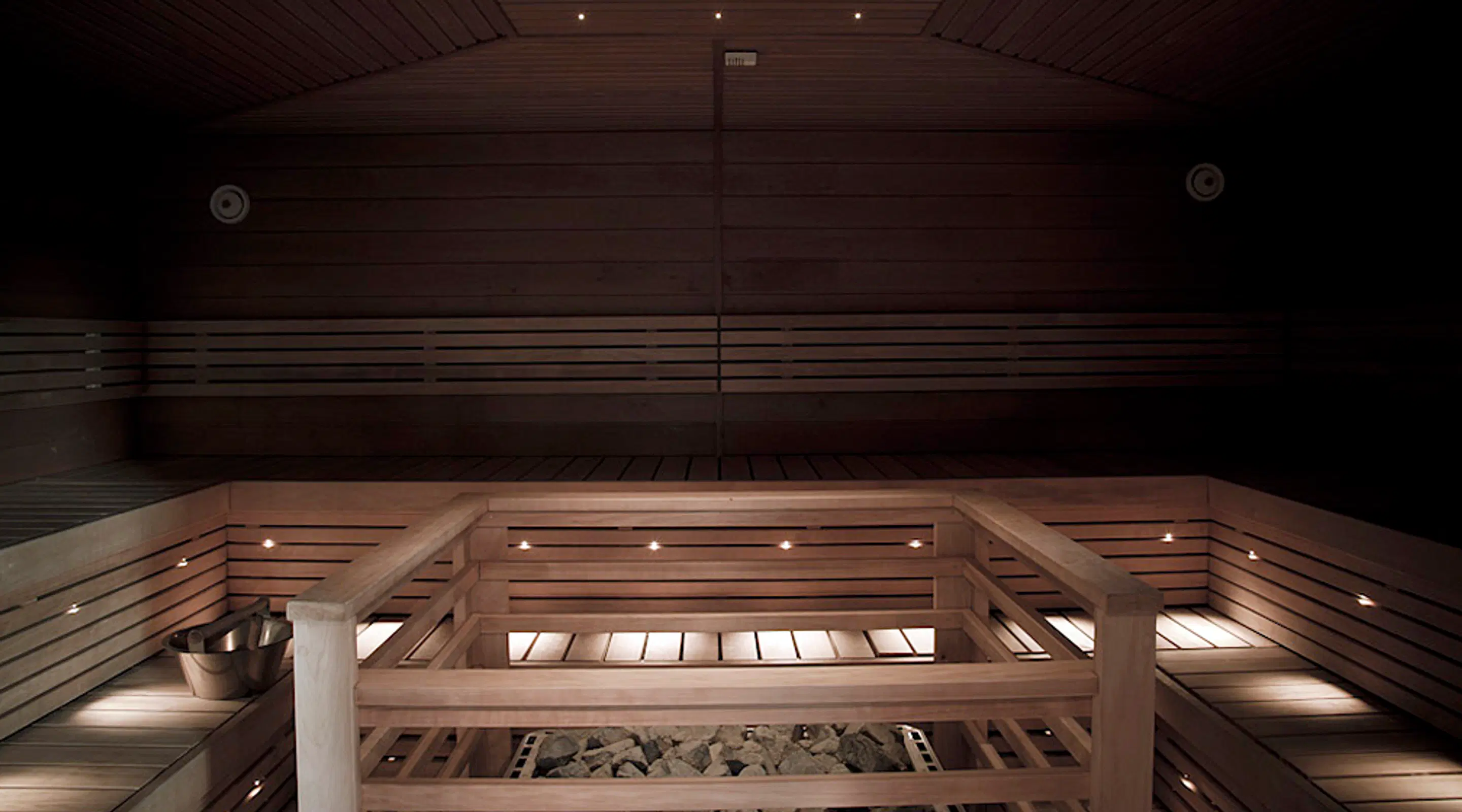 Dimly lit wooden sauna interior. Wooden benches and a heater with sauna stones visible in foreground. Minimalist lighting creates atmosphere.