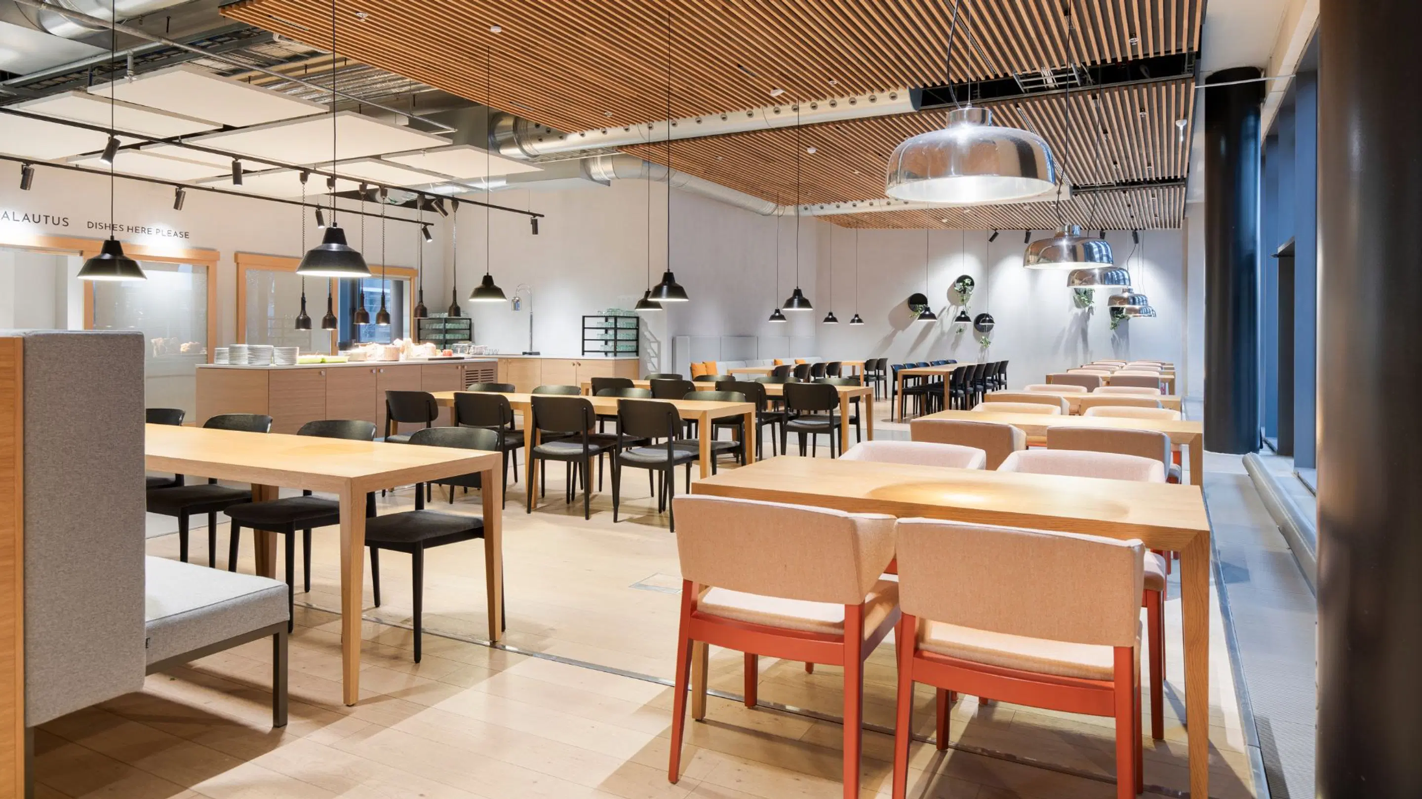 Modern, ljus restaurang med träbord, svarta och orangea stolar samt träribbor i taket. Öppet utrymme med jämnt golv och god belysning.