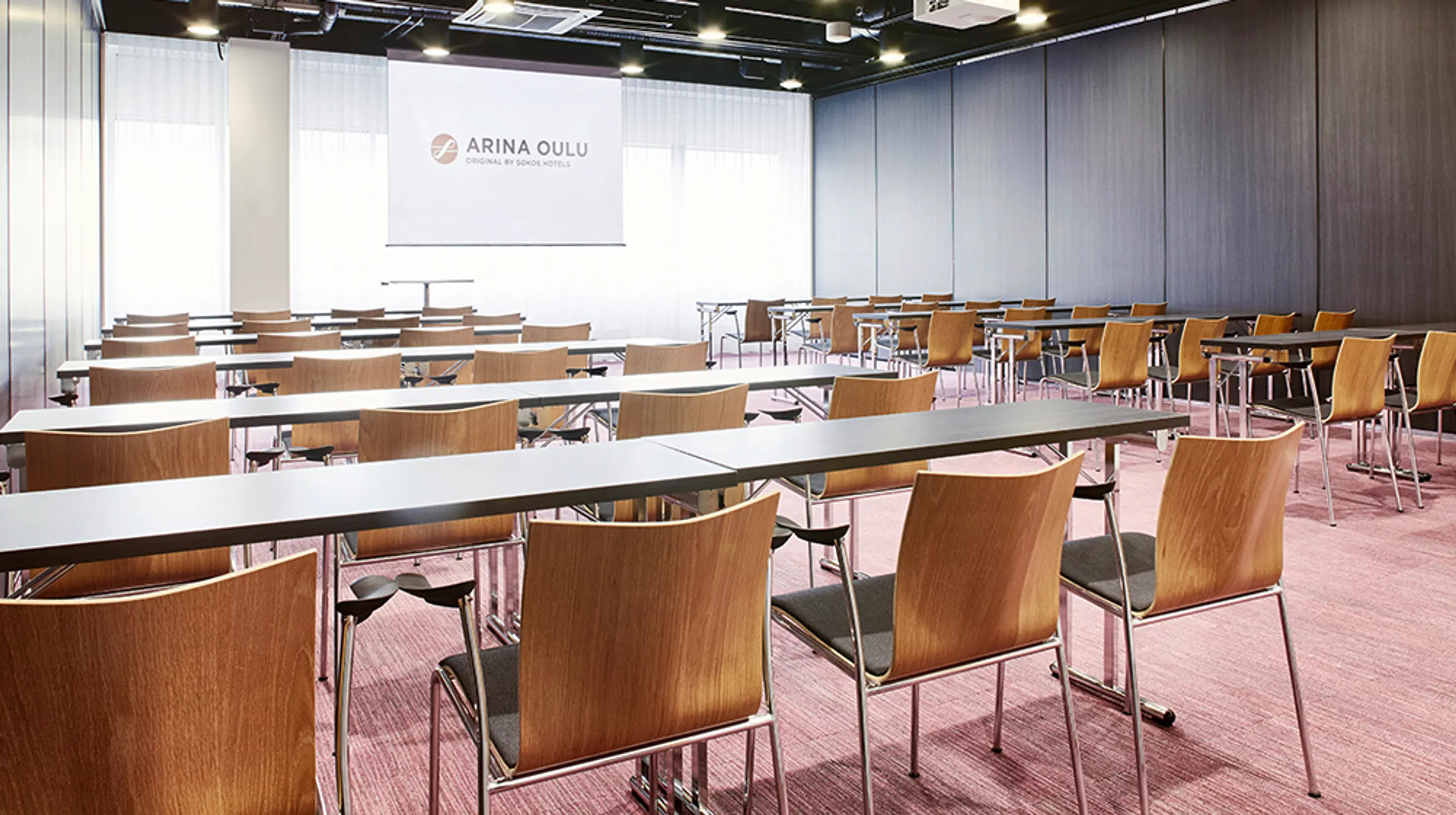Conference room with rows of white tables and wooden-backed chairs. The room features wood panel walls and a pink wall-to-wall carpet.