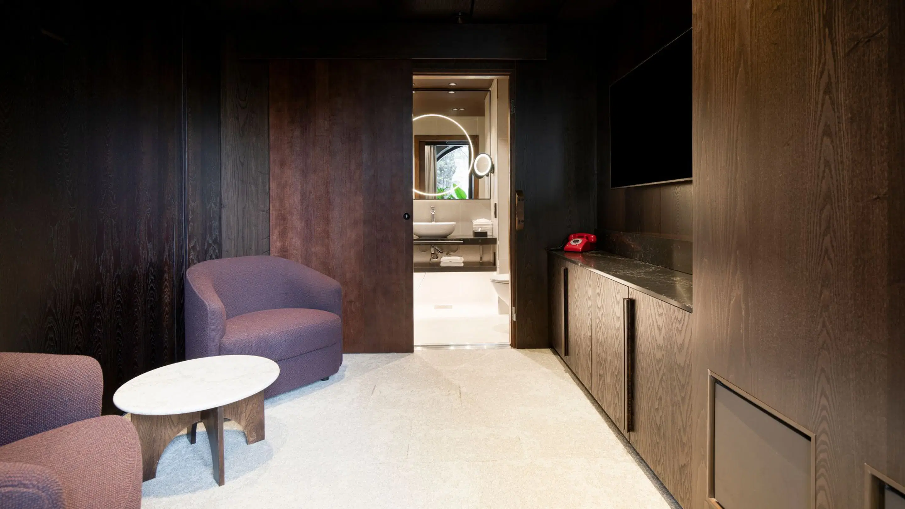Stylishly decorated hotel room with dark wood paneling, purple armchair, light-colored floor, and view into bathroom featuring circular illuminated mirror.