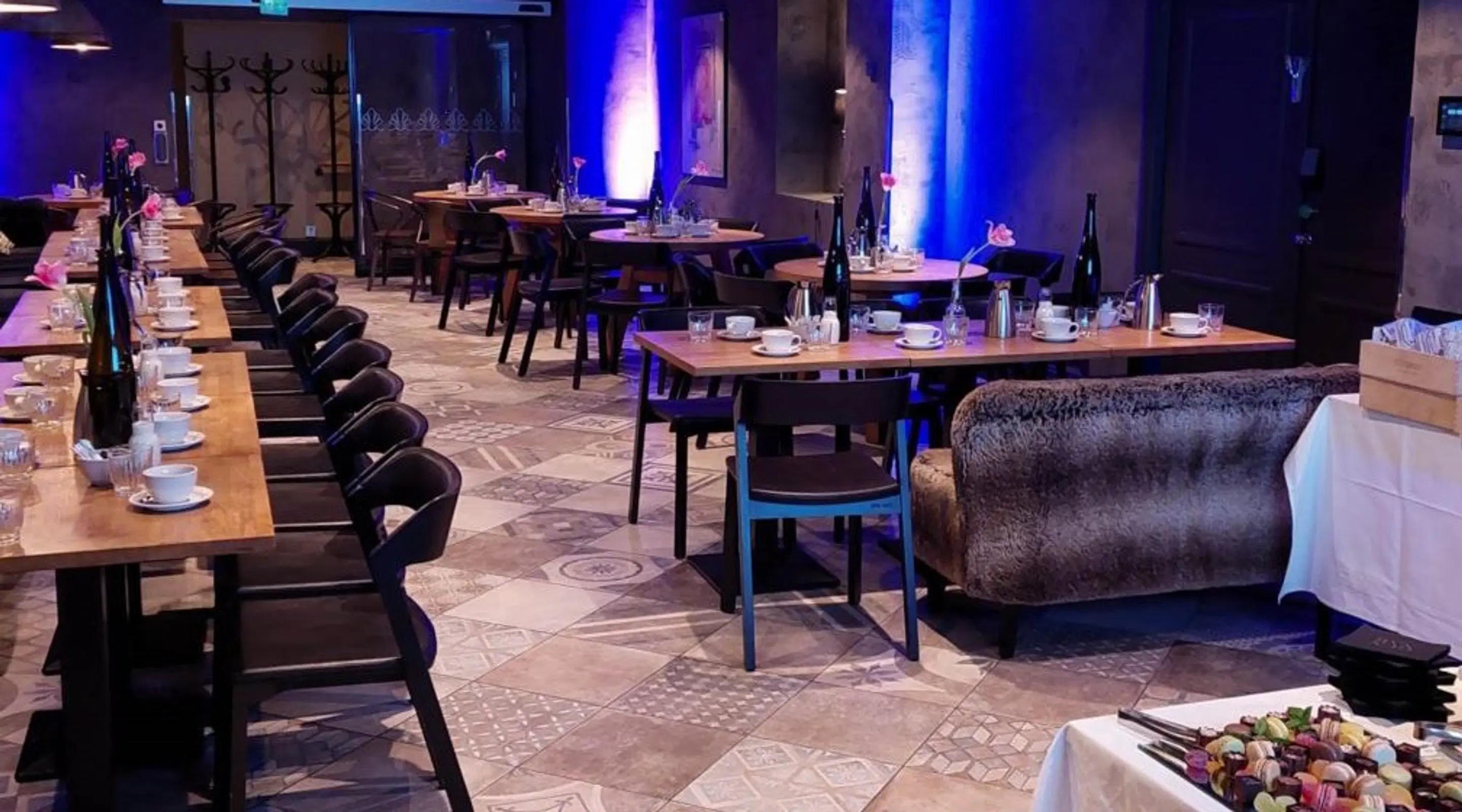 Restauranginredning med blå belysning. Flera träbord dukade med porslin, ett brett mönstrat klinkergolv och en soffhörna.