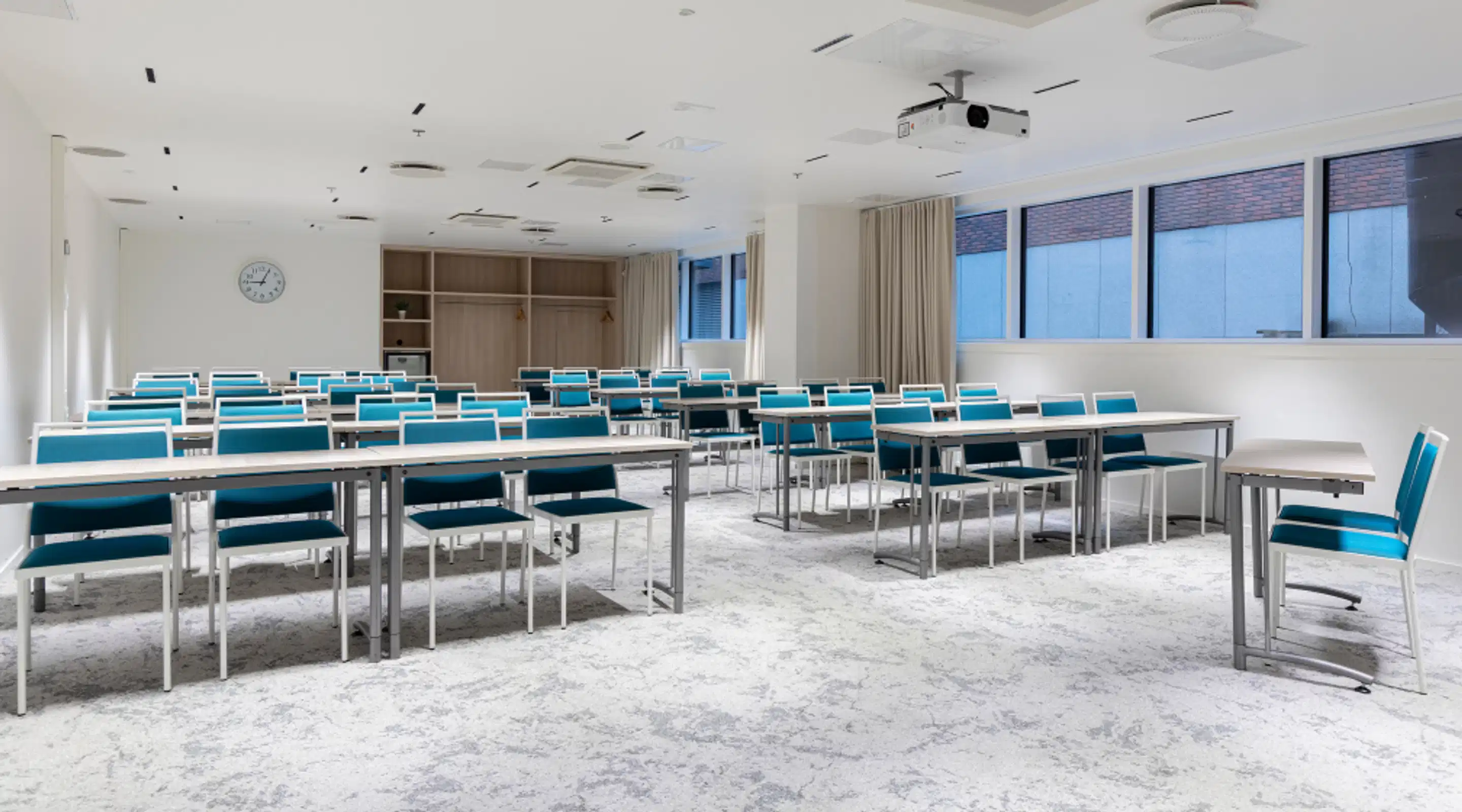 Bright meeting room with blue chairs and white tables arranged in rows. Wall clock and ceiling-mounted projector. Light gray carpet floor.