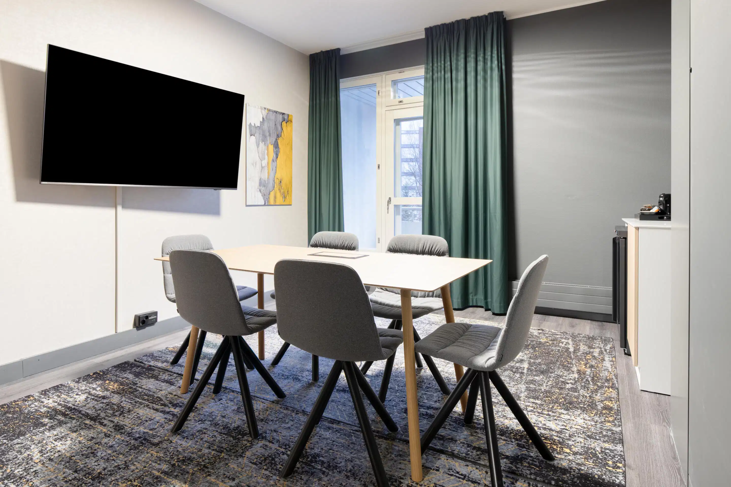 Modern meeting room with white table, six gray chairs, wall-mounted television, and green curtains covering a window.