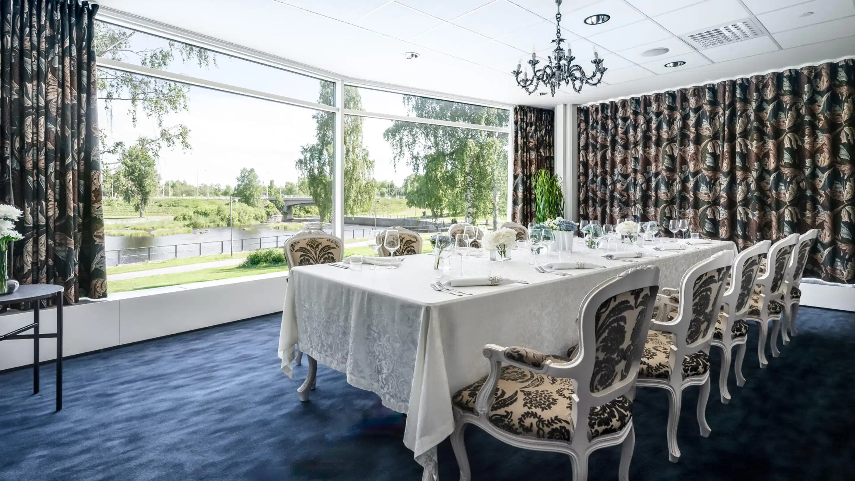 Elegant dining room with long table covered in white tablecloth. Ornate chairs, large windows overlooking a river and park landscape.